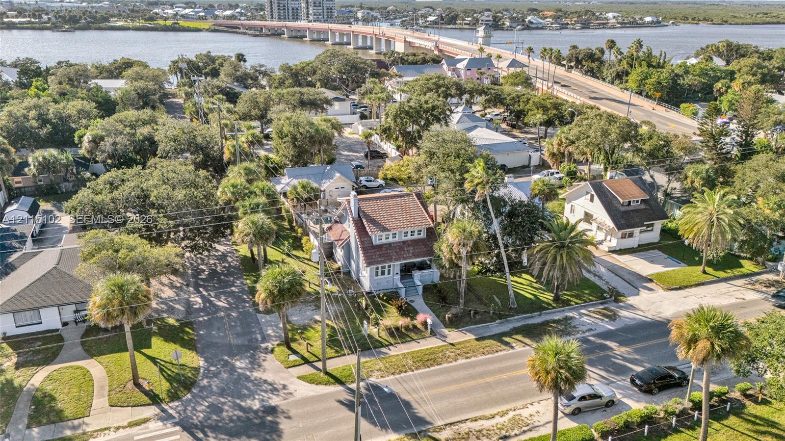 Aerial perspective of coastal residential property featuring mature palm landscaping, multi-story structure with a pitched roof, and paved street frontage