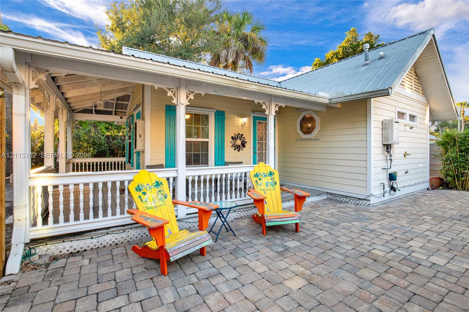 Covered wrap-around porch with decorative millwork, light-colored horizontal siding, metal roofing, and stone paver patio