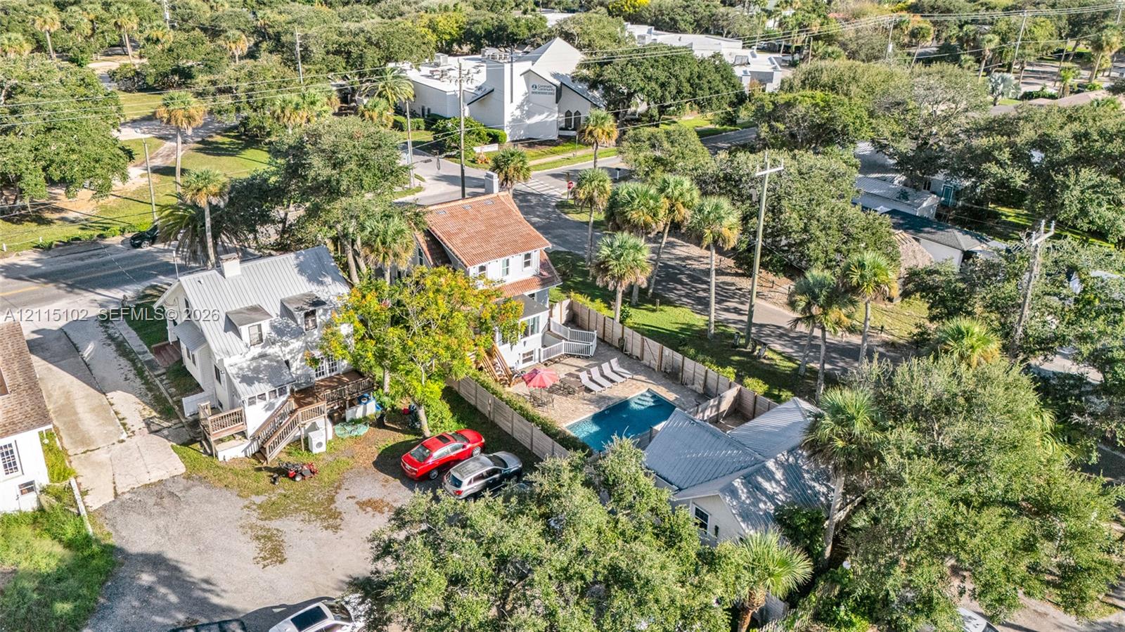 Aerial perspective featuring an in-ground swimming pool, wood fencing, multiple structures with metal roofing, and mature palm trees