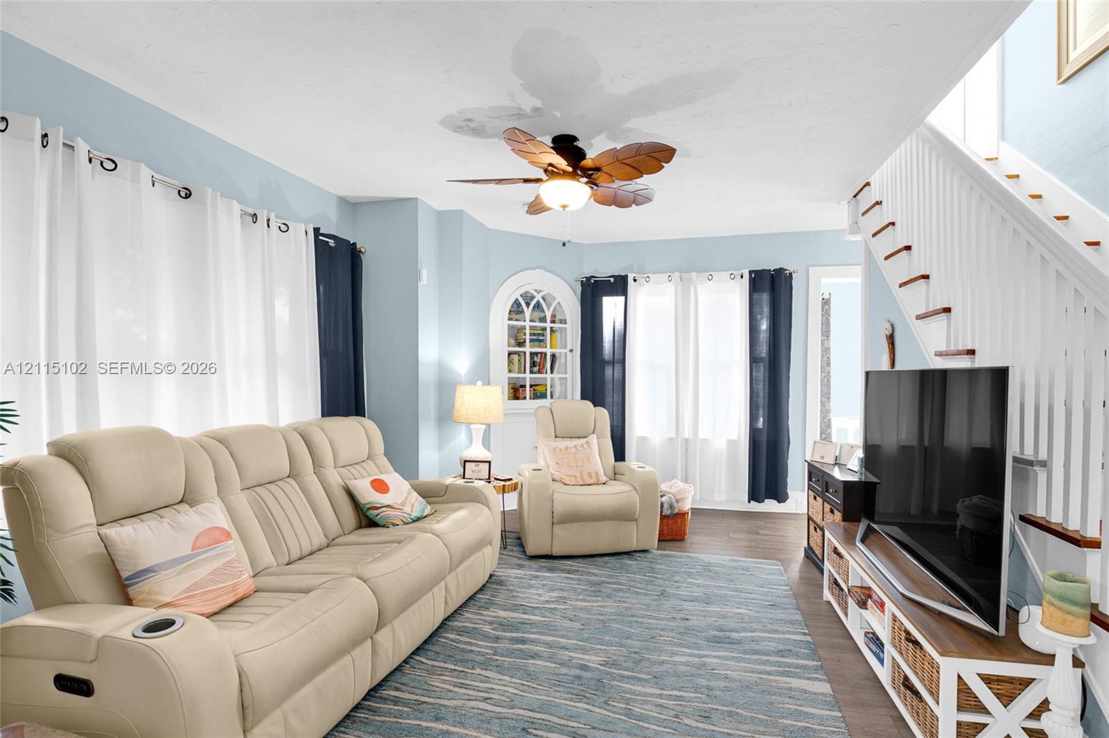 Living area featuring wood-finish flooring, a ceiling fan with leaf-shaped blades, arched built-in shelving, and a staircase with white balusters