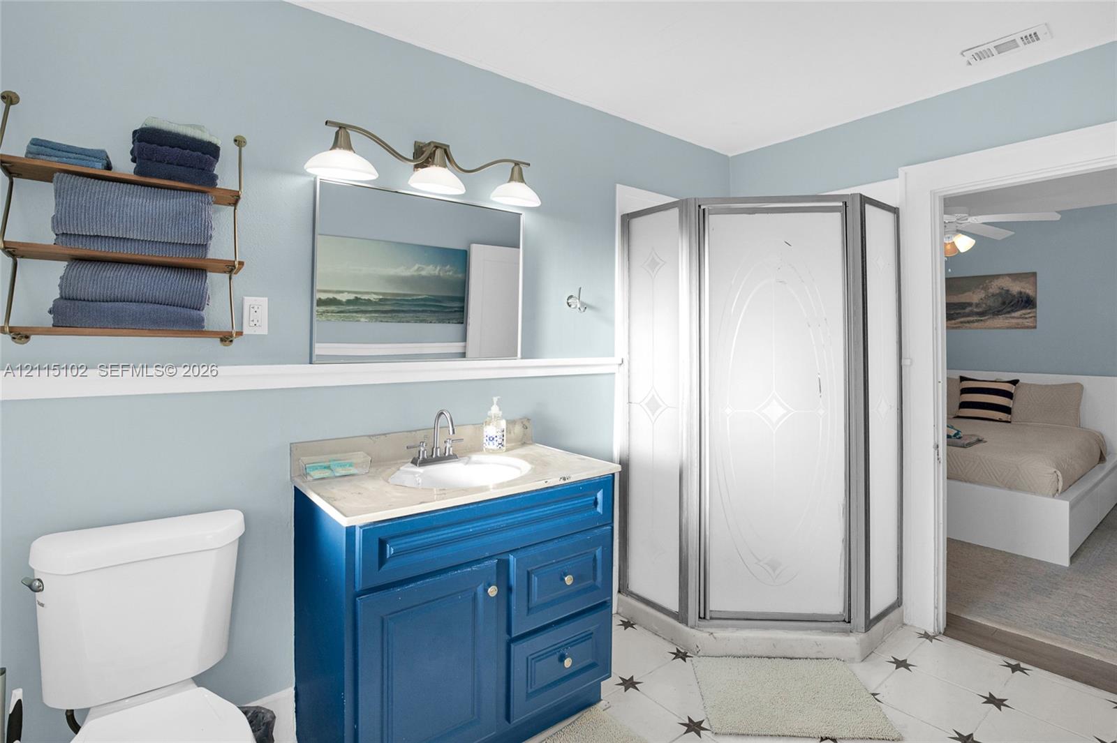 Bathroom featuring a vanity with integrated sink and stone-finish countertop, corner shower enclosure with patterned glass, wall-mounted shelving unit, and three-light fixture