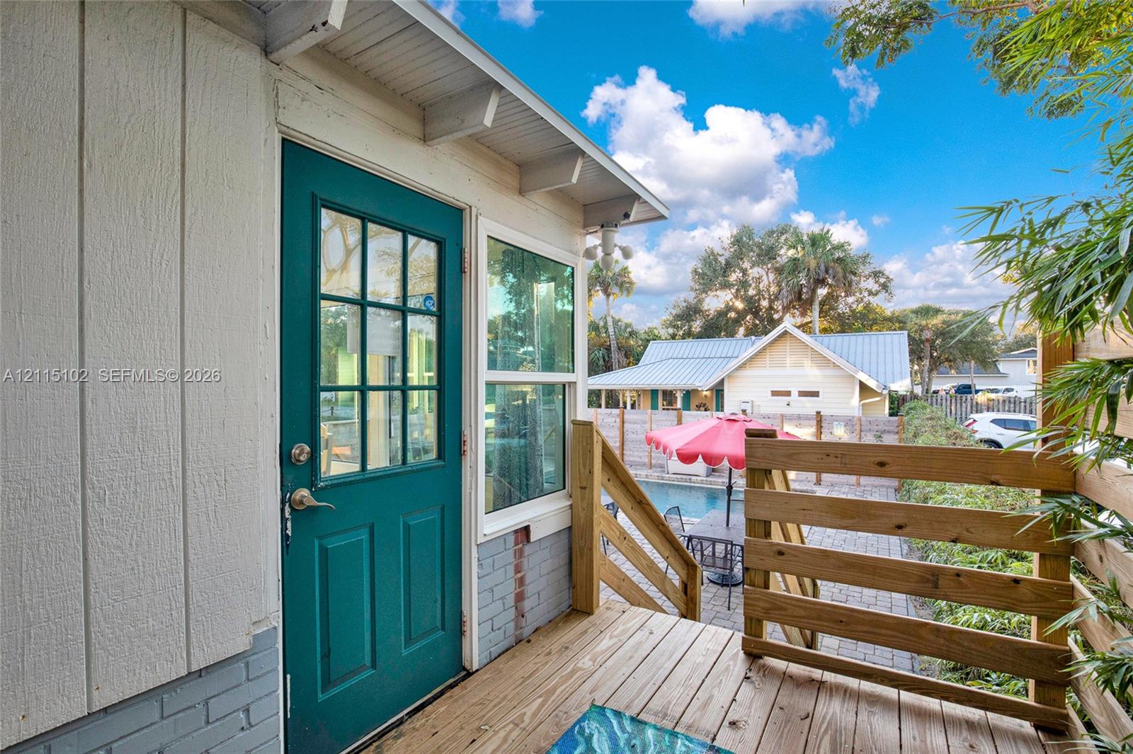 Exterior deck featuring horizontal wood railings, teal-painted door with grid window, paneled siding, and a brick base