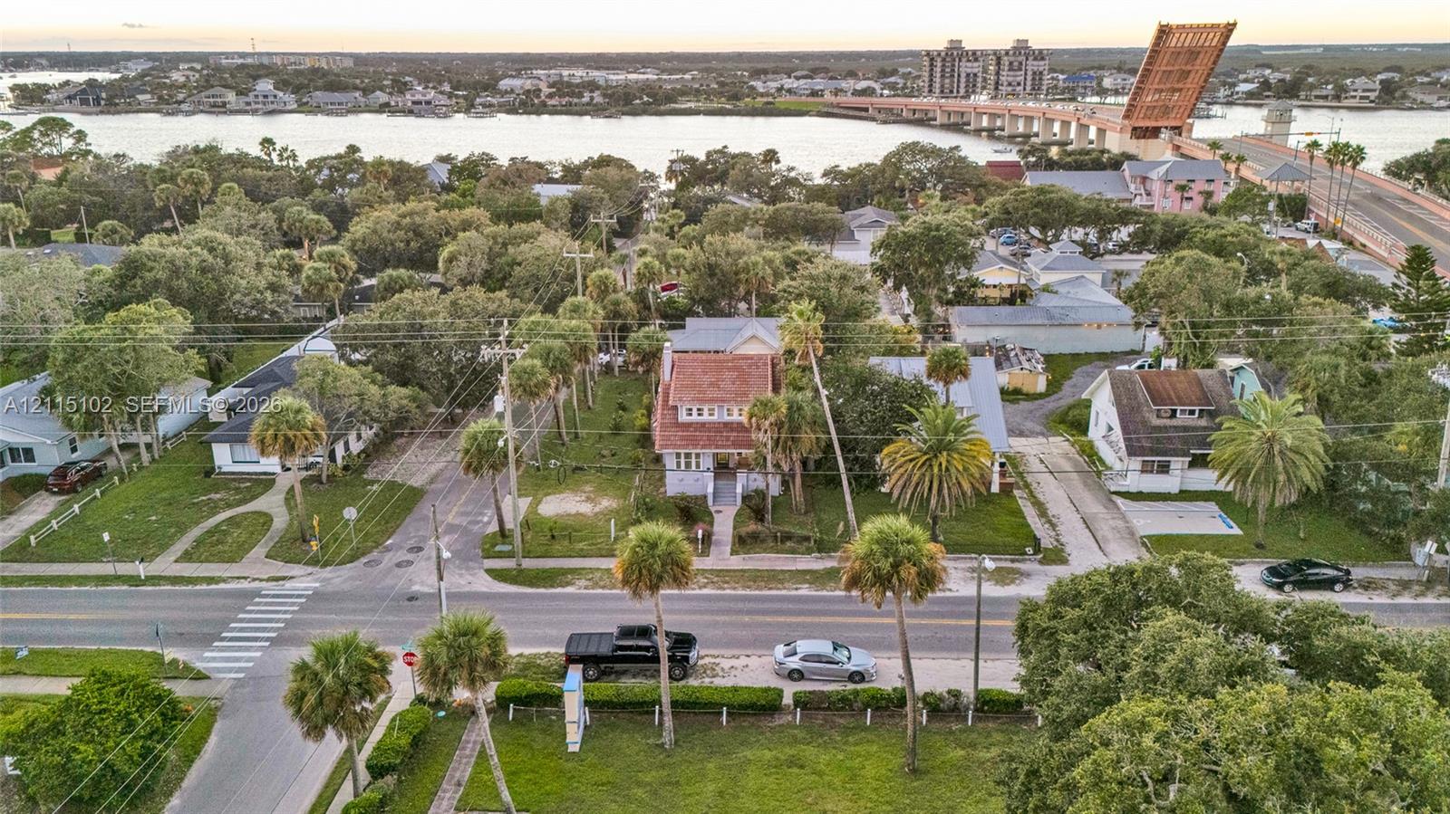 Aerial perspective featuring a multi-story residence with a pitched tile roof, situated in a coastal area with mature palms and proximate to a waterway and bridge