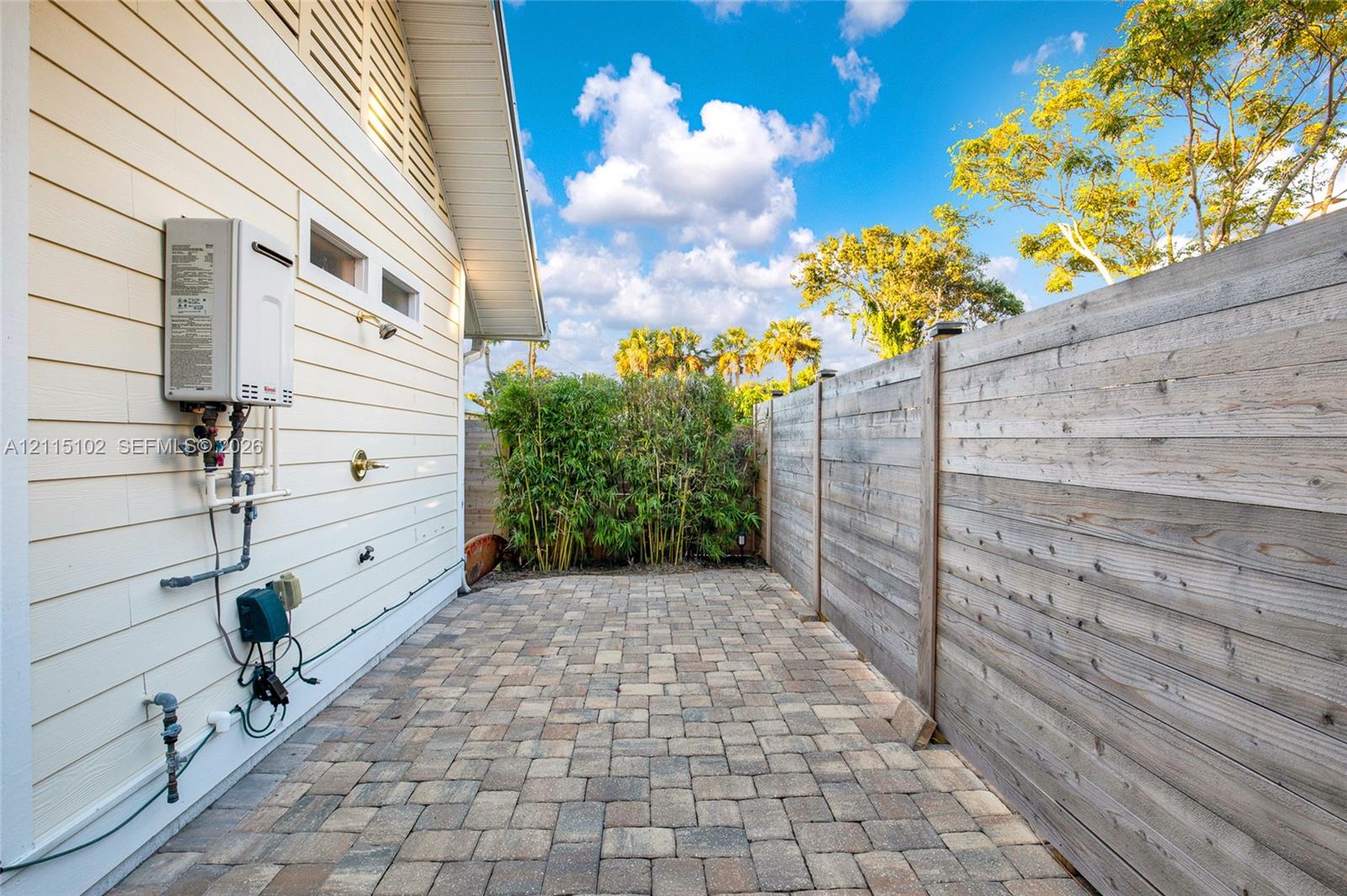 Exterior side patio featuring light-colored horizontal lap siding, brick paver flooring, a wall-mounted tankless water heater, an outdoor shower fixture, and a wood privacy fence