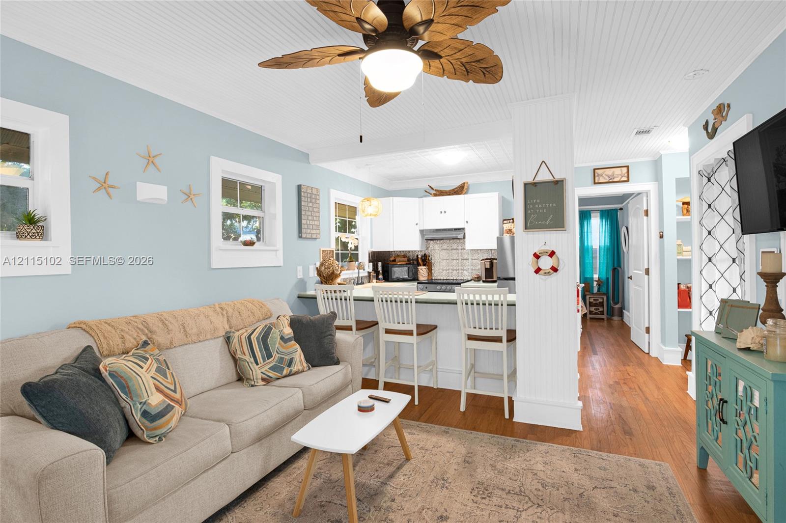 Open-concept living space featuring wood-finish flooring, beadboard ceiling detail, integrated breakfast bar with white cabinetry, and palm-leaf ceiling fan