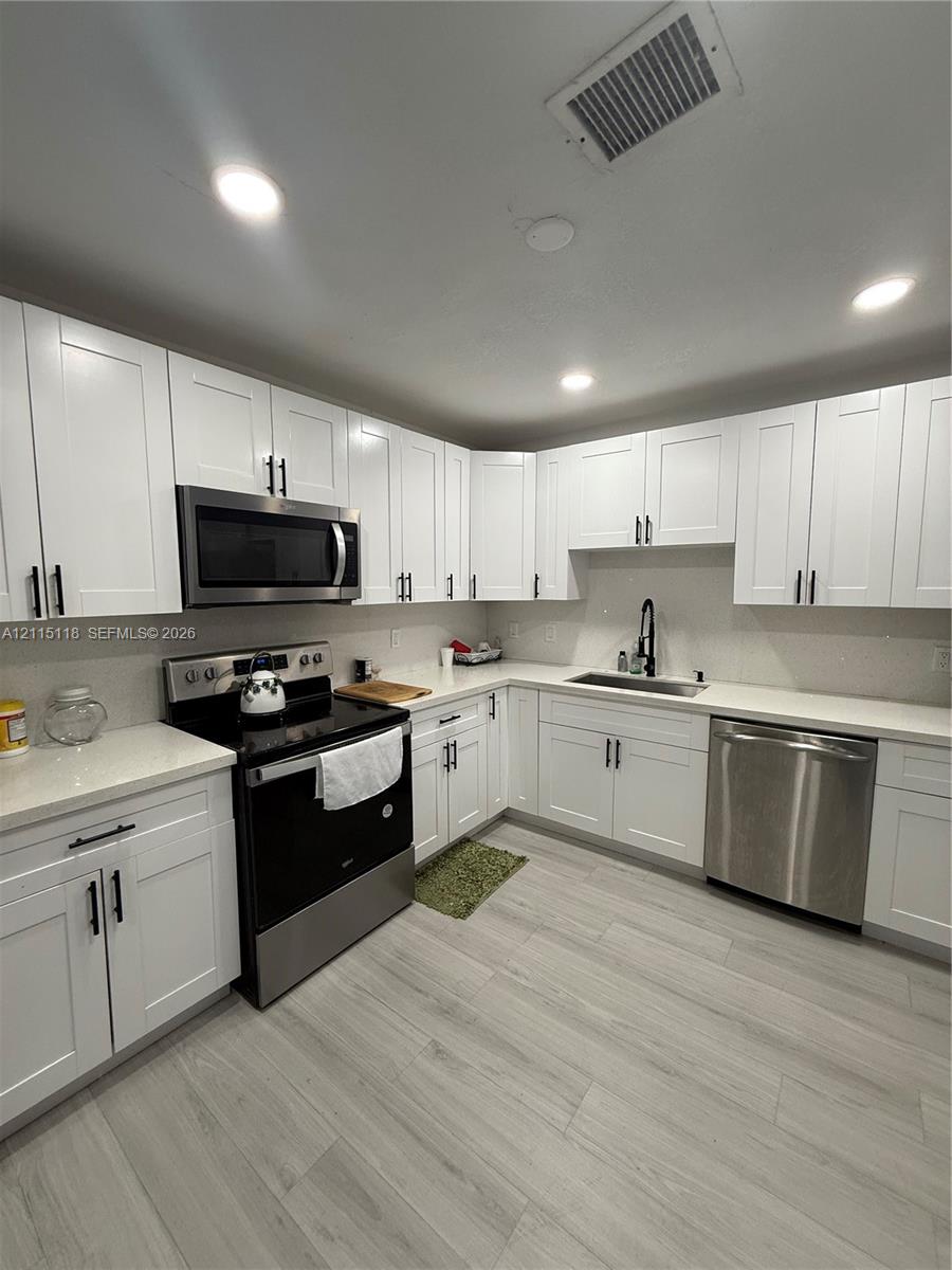 Kitchen featuring white Shaker-style cabinetry, stainless steel appliances, light-toned countertops, a stainless steel undermount sink with a matte black faucet, and wood-finish flooring