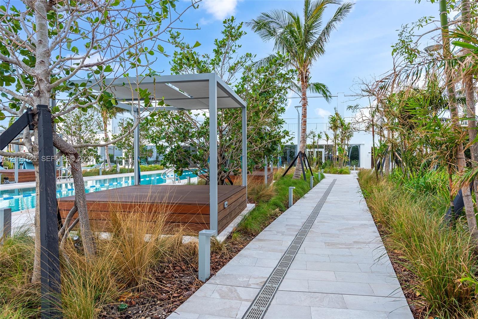 Outdoor lounge area featuring a wood-deck platform with a modern metal-frame canopy, stone paver pathway, and mature tropical landscaping