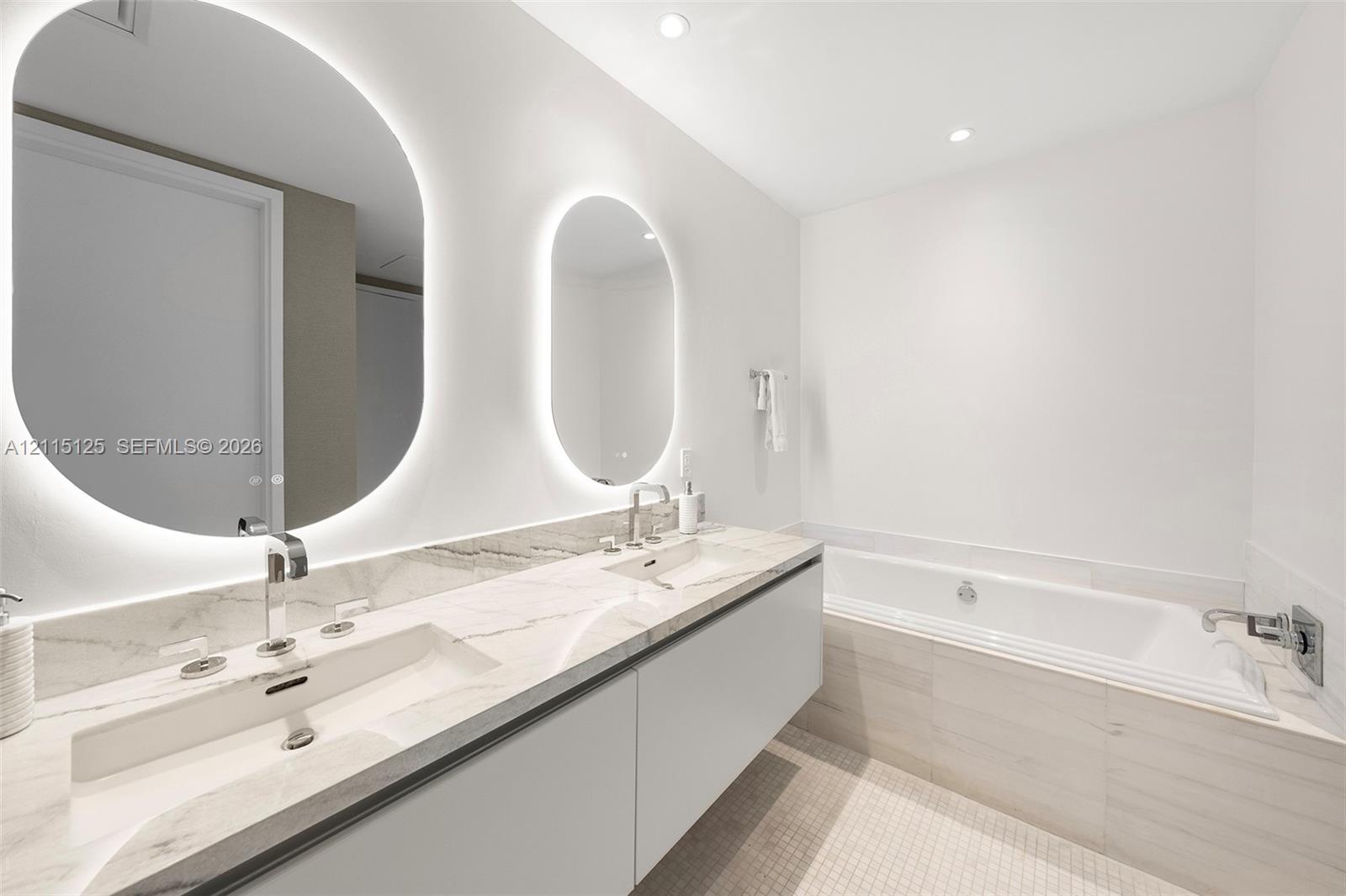 Floating dual-sink vanity with a solid surface countertop, backlit oval mirrors, and a deep soaking tub featuring tiled surround