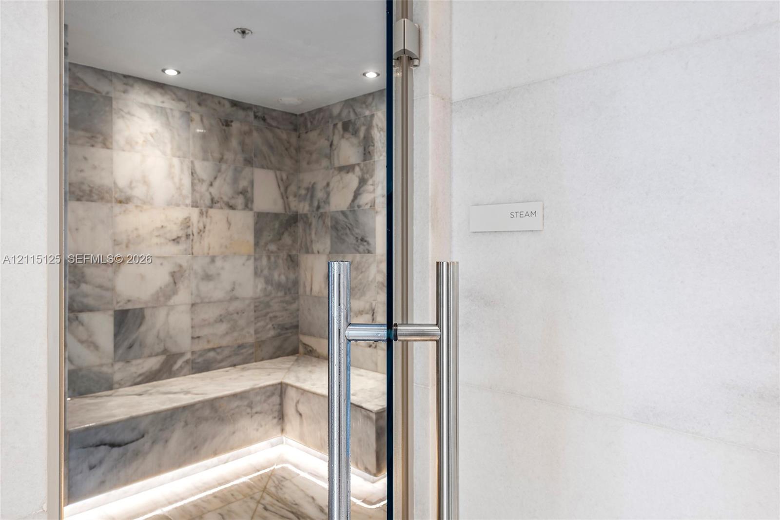 Steam room featuring stone-tiled walls, built-in stone bench seating, recessed lighting, and a frameless glass door with a stainless steel handle