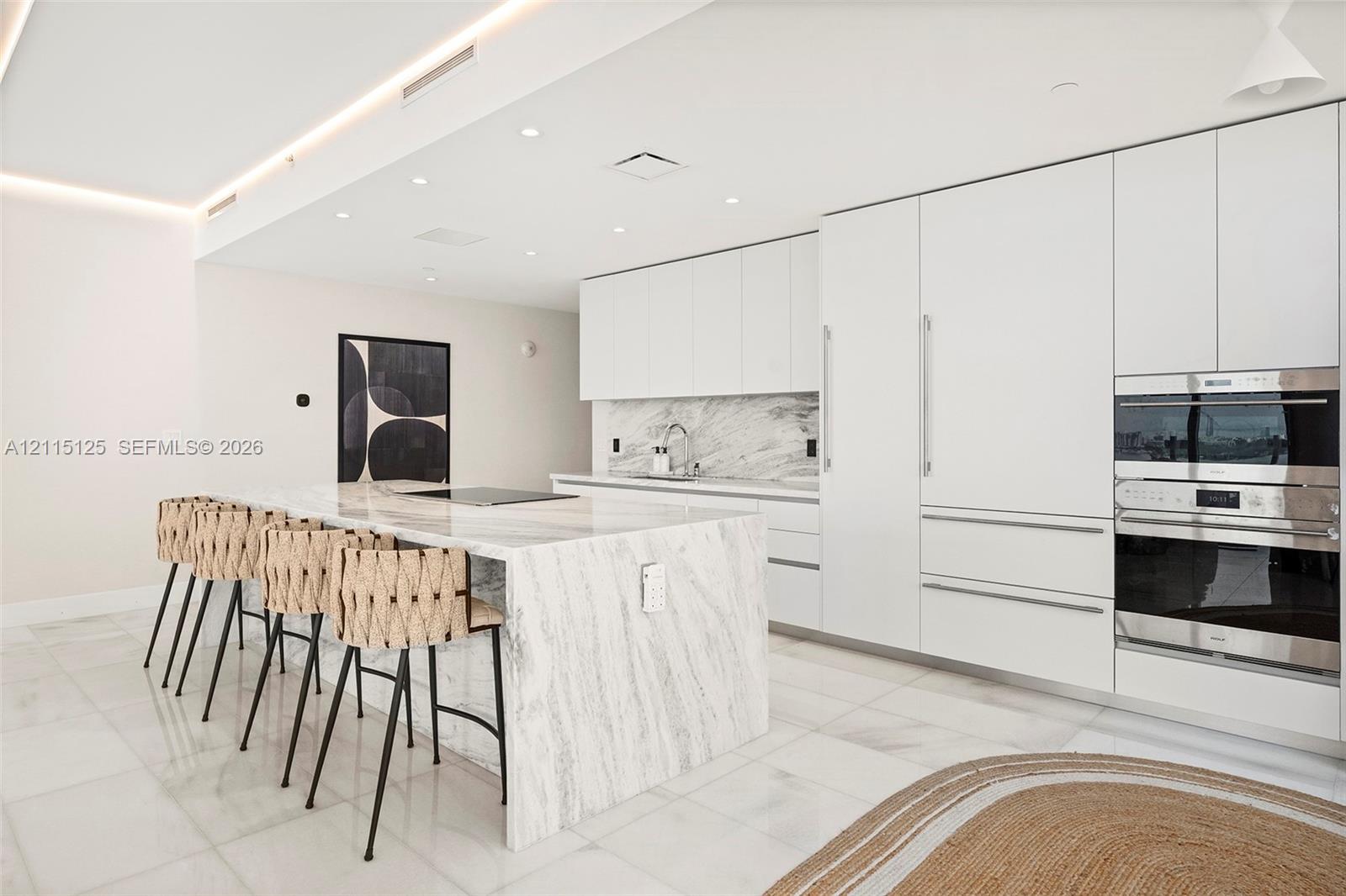 Kitchen featuring a waterfall island with stone countertop, floor-to-ceiling cabinetry, integrated stainless steel double wall ovens, recessed ceiling lighting, and large-format tile flooring
