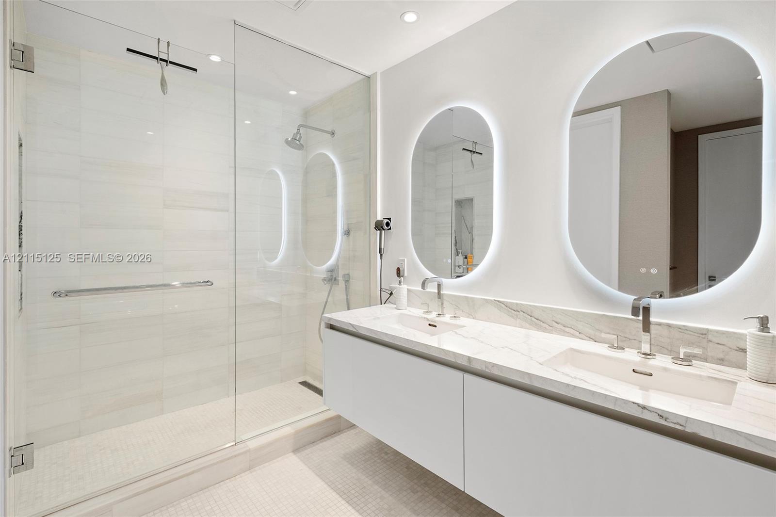 Floating dual vanity with stone countertop, integrated sinks, chrome fixtures, backlit oval mirrors, and a glass-enclosed shower featuring tile surrounds
