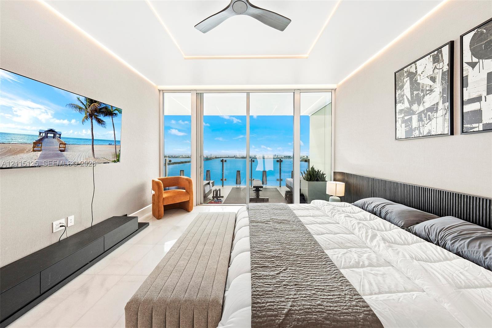 Floor-to-ceiling glass sliders, tray ceiling with recessed perimeter lighting, modern ceiling fan, dark slatted wood headboard, and polished stone flooring