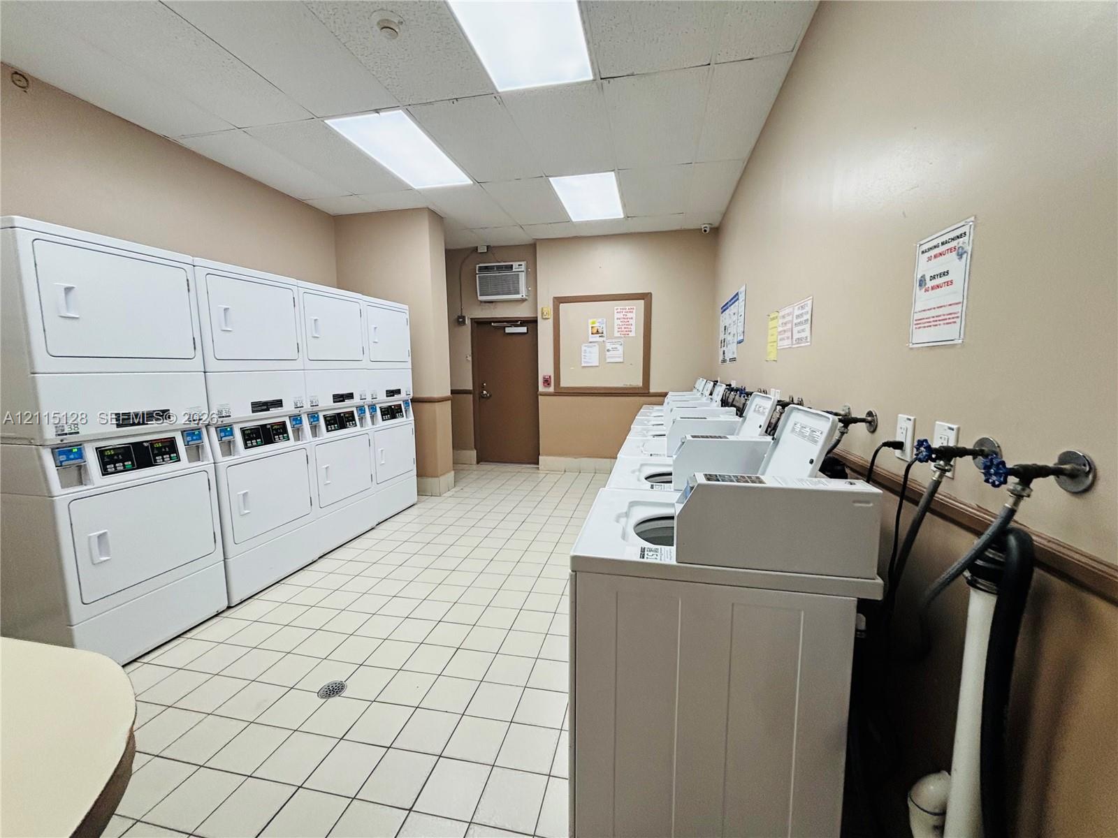 Laundry facility featuring multiple stacked washer-dryer units, high-capacity top-loading washing machines, ceramic tile flooring, and drop ceiling lighting