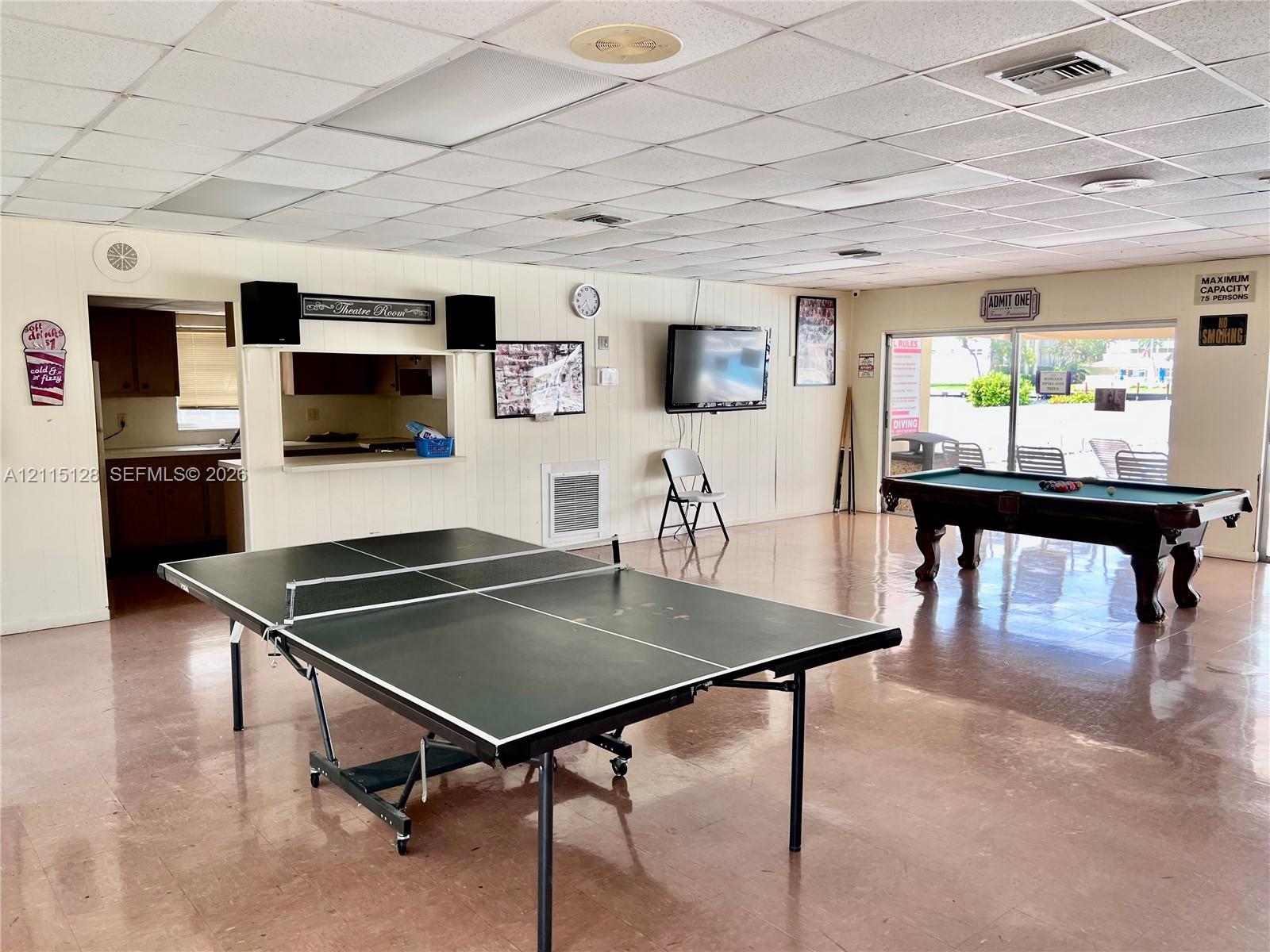 Open-concept recreational space featuring panelled walls, tile flooring, a drop ceiling with recessed lighting, and an integrated kitchenette pass-through window