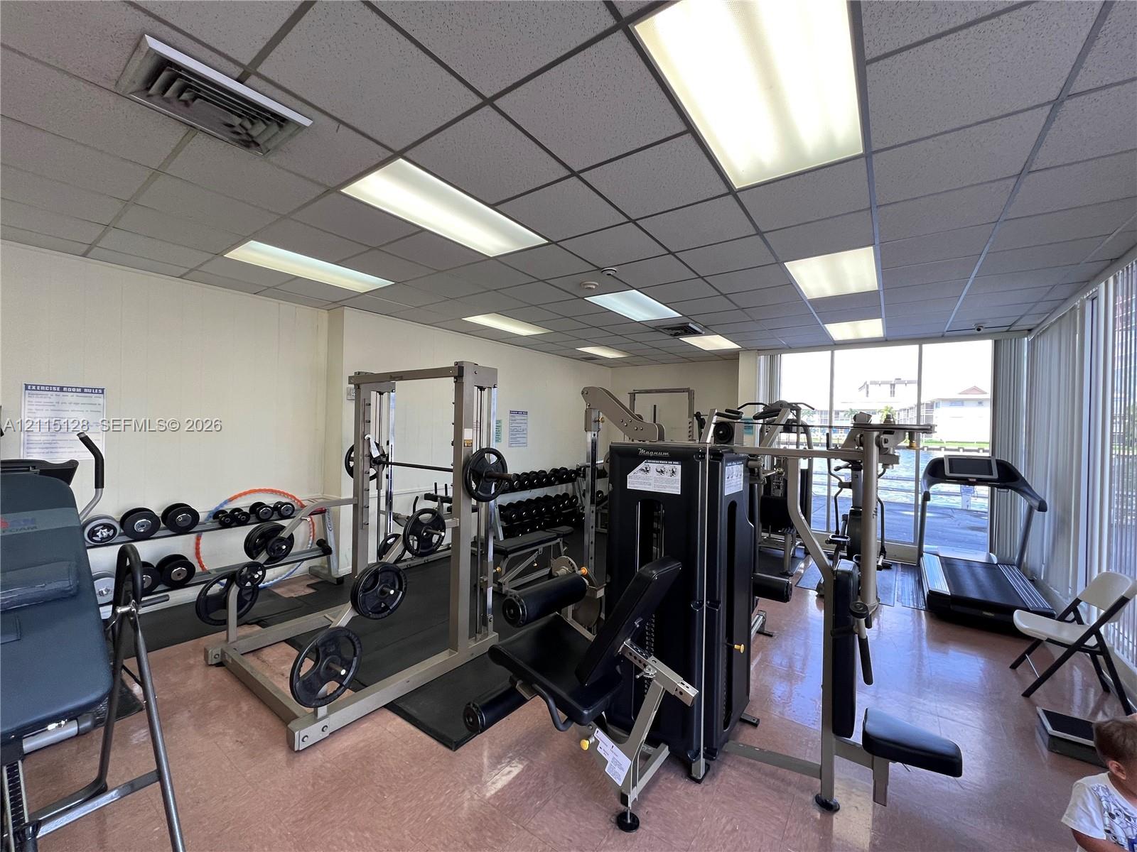 Fitness room featuring floor-to-ceiling windows, drop-ceiling grid with recessed lighting, and vinyl composition tile flooring Fitness room featuring floor-to-ceiling windows, drop-ceiling grid with recessed lighting, and vinyl composition tile flooring