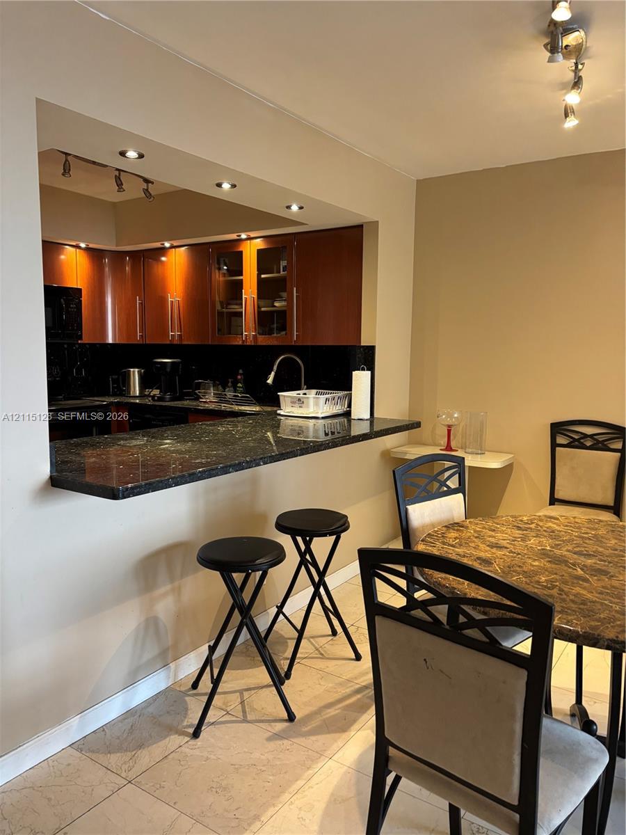 Kitchen featuring a stone-surfaced breakfast bar, wood cabinetry with glass-panel upper doors, integrated recessed lighting, and tile flooring Kitchen featuring a stone-surfaced breakfast bar, wood cabinetry with glass-panel upper doors, integrated recessed lighting, and tile flooring