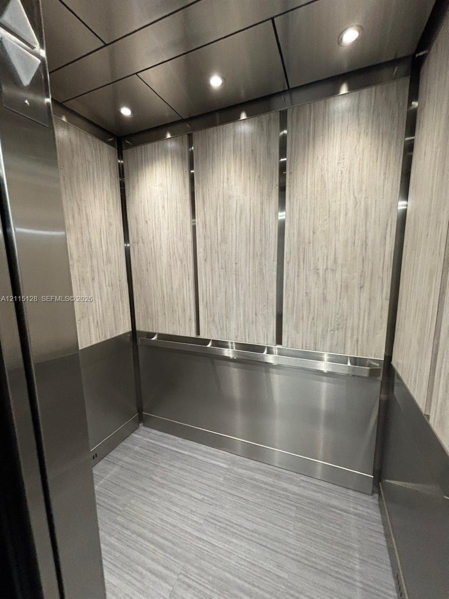 Elevator cab featuring vertical wood-finish wall paneling, stainless steel wainscoting and trim, recessed ceiling lighting, and tile flooring