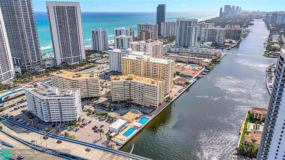 Aerial perspective of a high-rise residential complex featuring waterfront access, swimming pools, paved surface parking, and proximity to the Atlantic Ocean