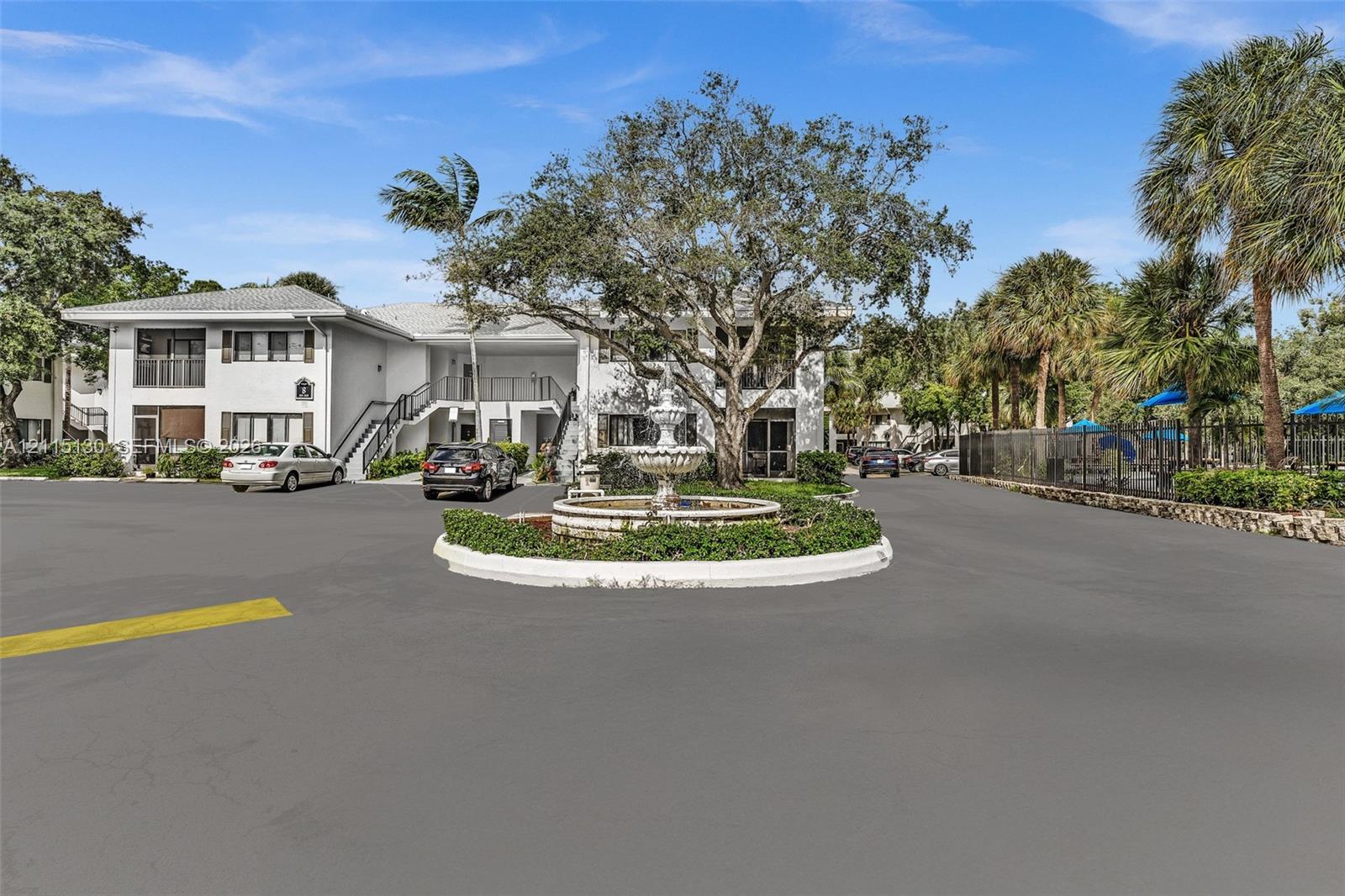 Exterior perspective featuring multi-story architecture, a central decorative stone fountain, manicured landscaping, and mature canopy trees