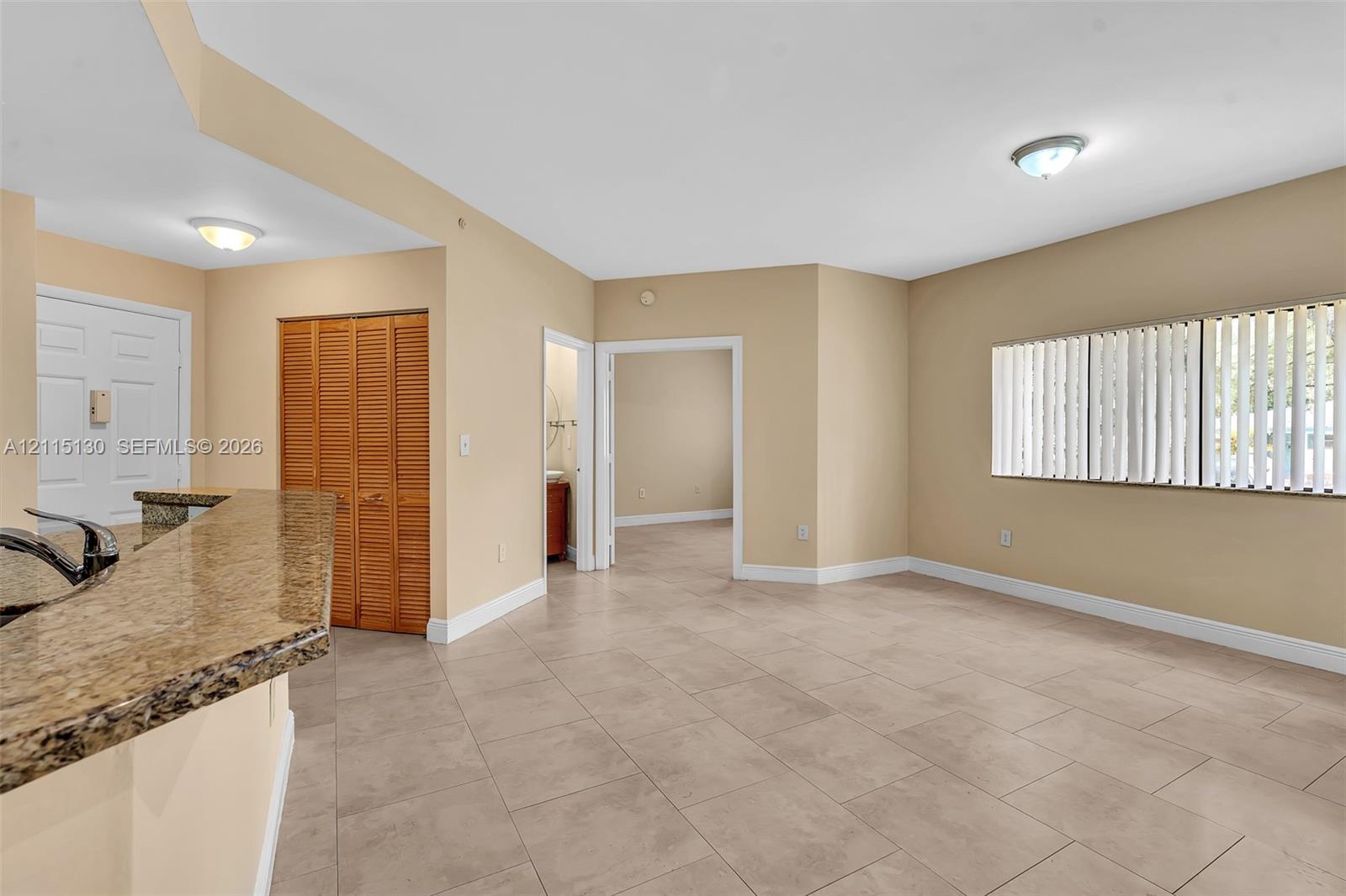 Open-concept interior featuring large-format ceramic tile flooring, kitchen breakfast bar with stone-finish countertop, dual flush-mount ceiling lights, and vertical blind window treatments