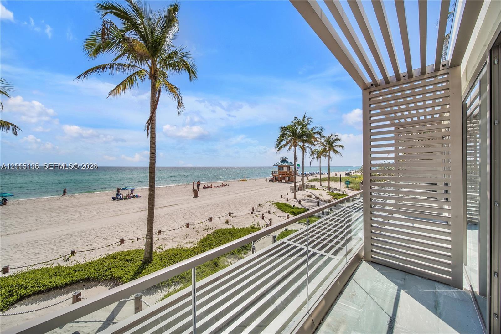 Private balcony featuring a glass panel railing, overhead slatted architectural screen, and stone flooring, oriented toward a coastal setting