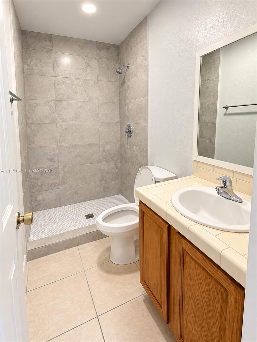 Bathroom featuring a walk-in shower with large-format wall tile and penny-tile flooring, wood-finish vanity with a tile countertop, recessed lighting, and a framed mirror