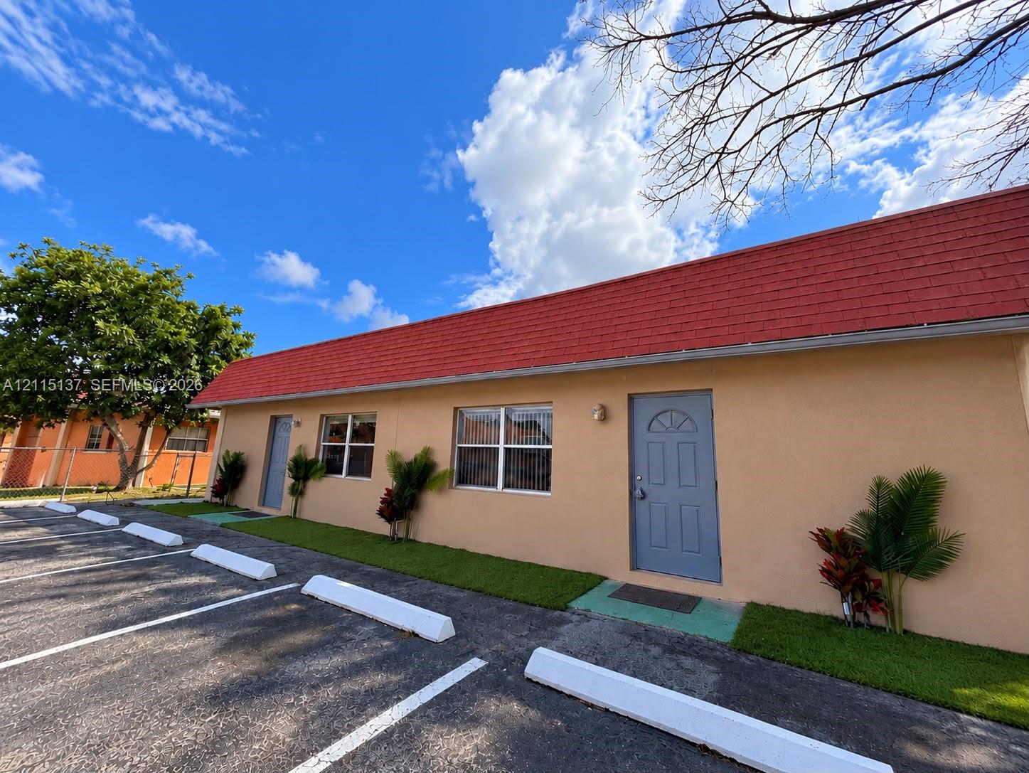 Single-story exterior featuring a red gable roof, stucco siding, and designated asphalt parking area with concrete bumpers
