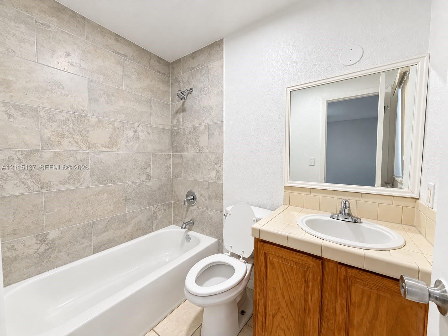 Bathroom featuring a tub-shower combination with large-format wall tile, wood-finish vanity cabinet with tile countertop, undermount sink, and framed wall-mounted mirror Bathroom featuring a tub-shower combination with large-format wall tile, wood-finish vanity cabinet with tile countertop, undermount sink, and framed wall-mounted mirror