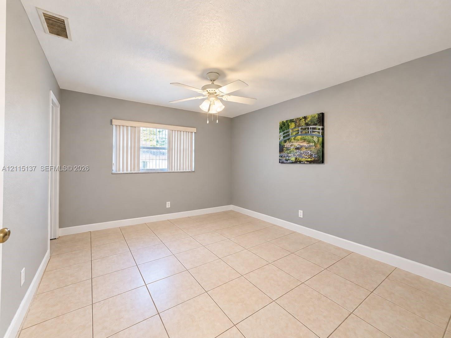 Interior room featuring large-format neutral floor tile, light grey wall paint, a multi-blade ceiling fan with an integrated light fixture, and a window with vertical blinds Interior room featuring large-format neutral floor tile, light grey wall paint, a multi-blade ceiling fan with an integrated light fixture, and a window with vertical blinds