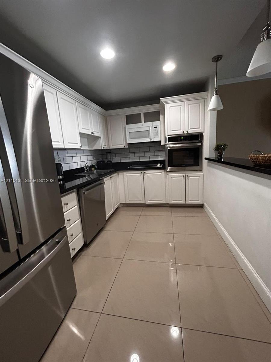 Kitchen featuring white shaker-style cabinetry, dark stone countertops, subway tile backsplash, stainless steel appliances including a built-in wall oven, and large-format tile flooring