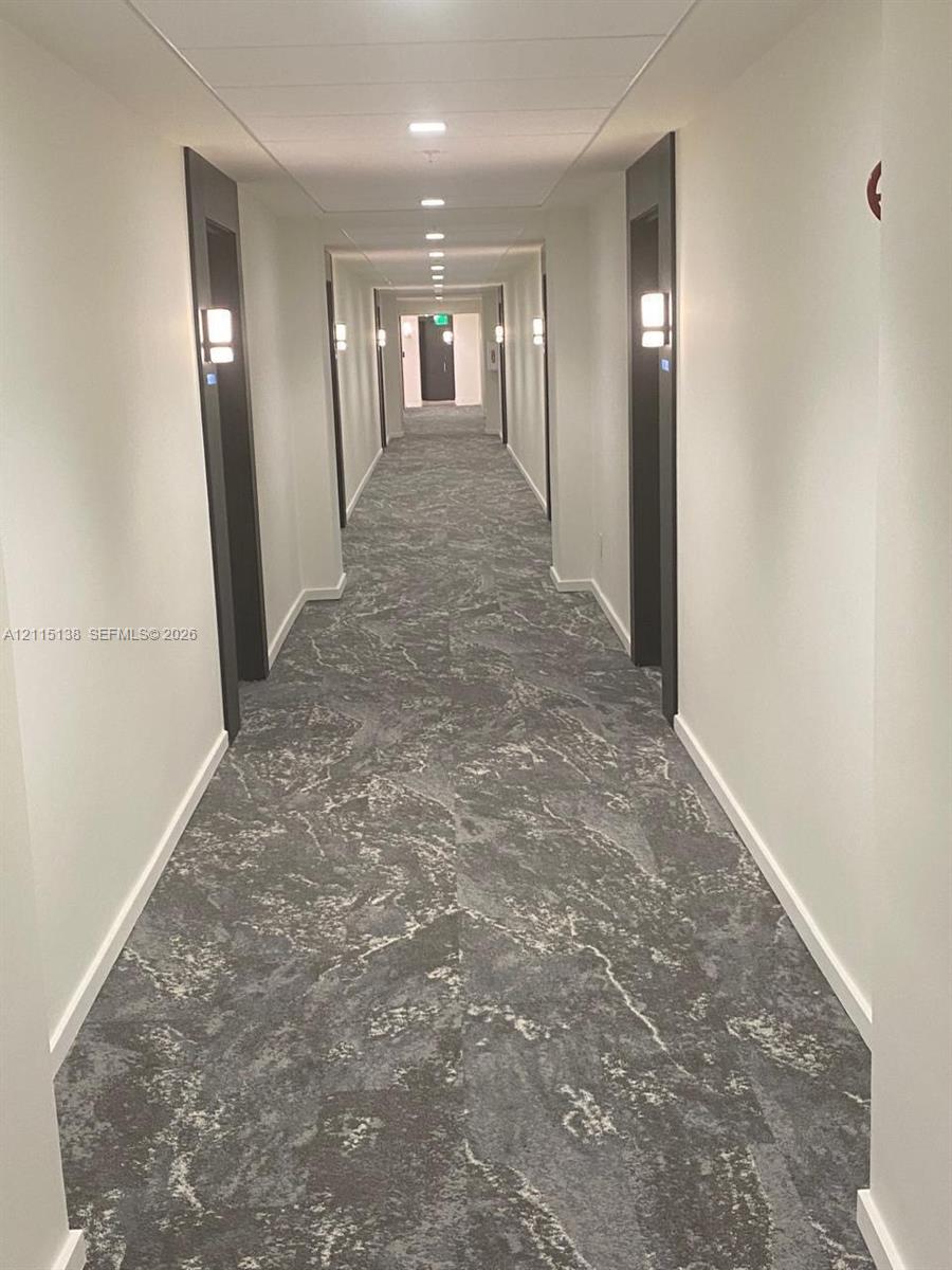 Hallway featuring patterned carpet flooring, recessed ceiling lighting, and wall-mounted light fixtures with dark door frames