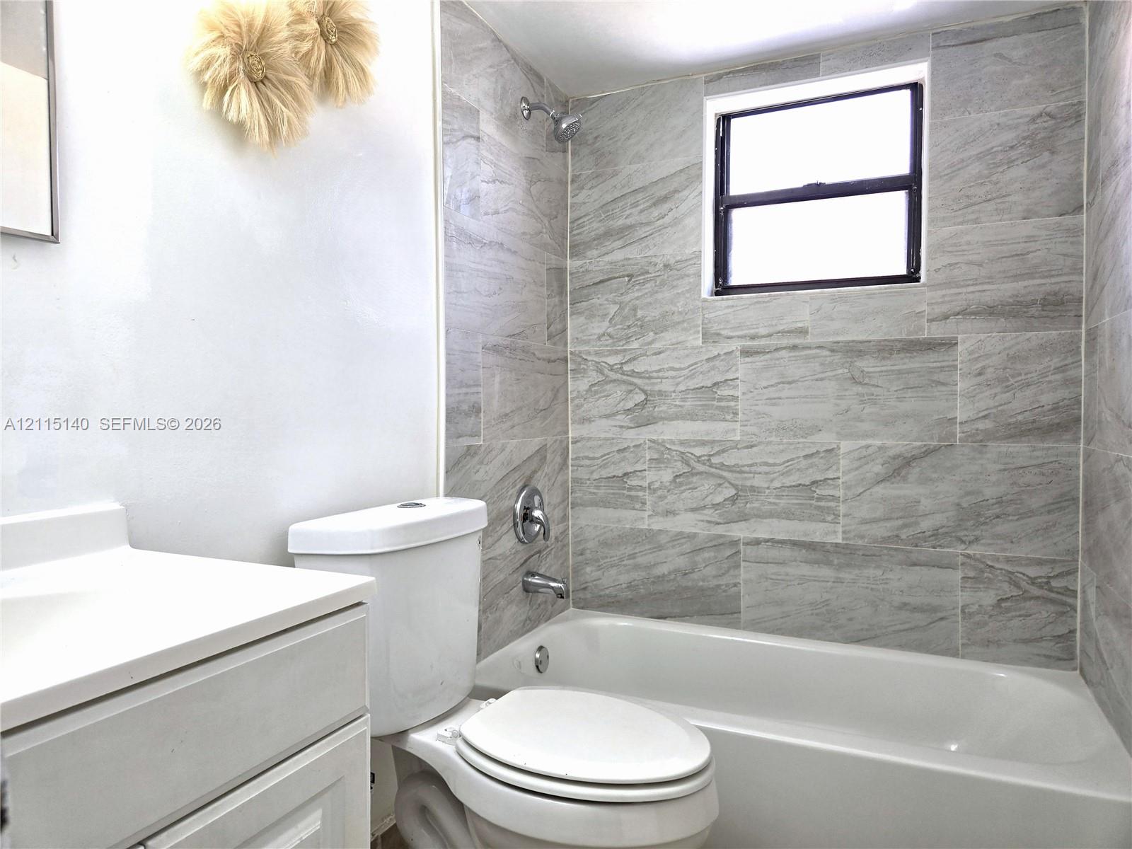 Bathroom featuring a shower-tub combination with grey-toned tile surround, a white vanity with an integrated sink, a built-in toilet, and an operable window
