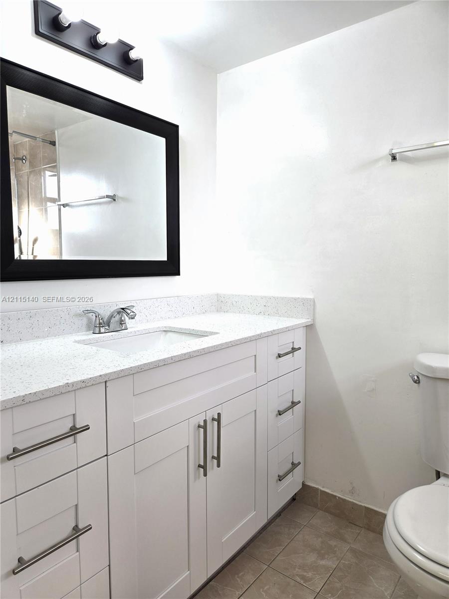 White shaker-style cabinetry with satin nickel pulls, stone-composite countertop with integrated sink, rectangular mirror with a black frame, three-light vanity fixture, and neutral tile flooring
