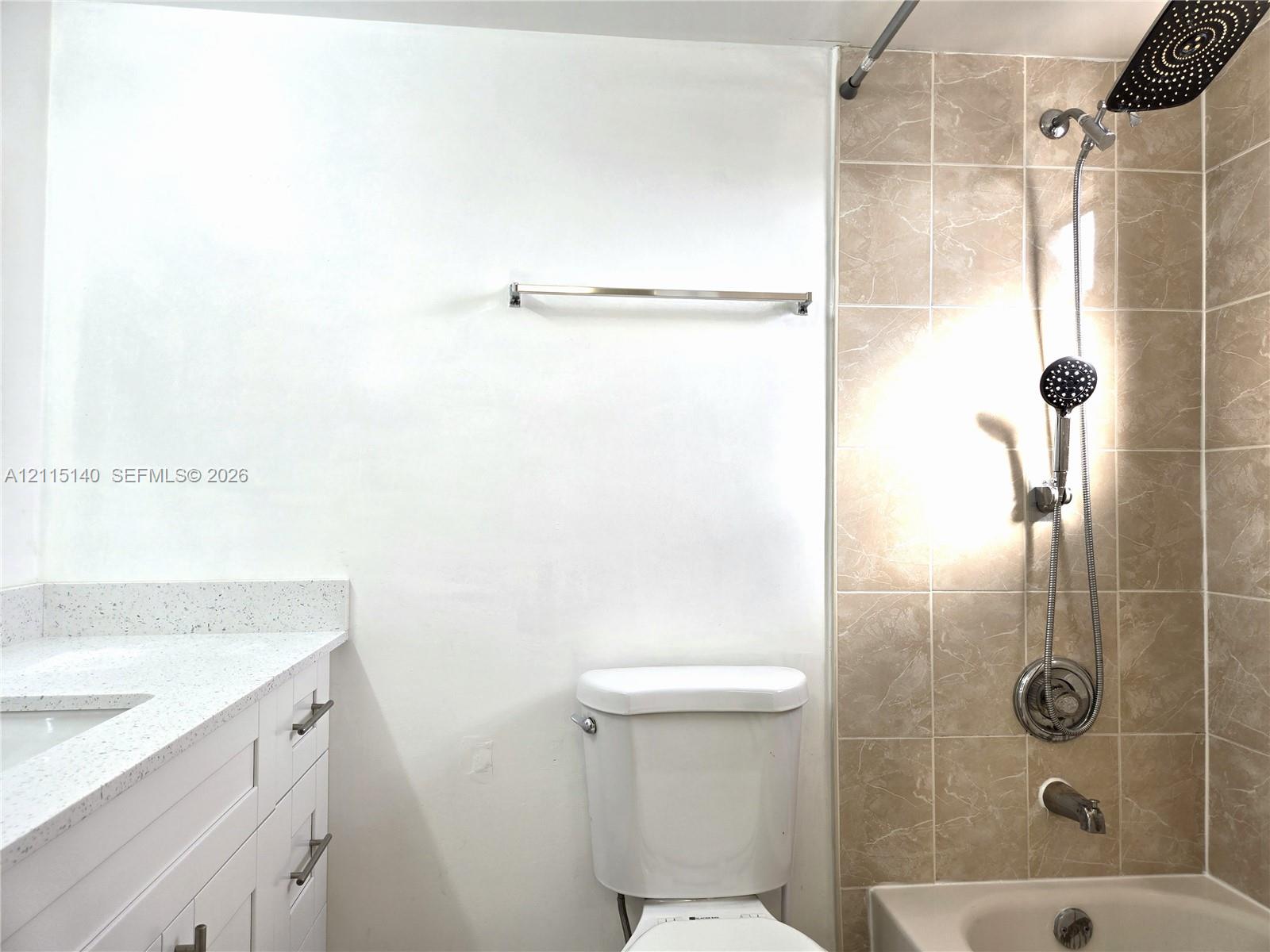 White vanity with light-speckled countertop and rectangular basin, ceramic tile shower surround, stainless steel rainfall showerhead with handheld attachment, and wall-mounted metal towel rack