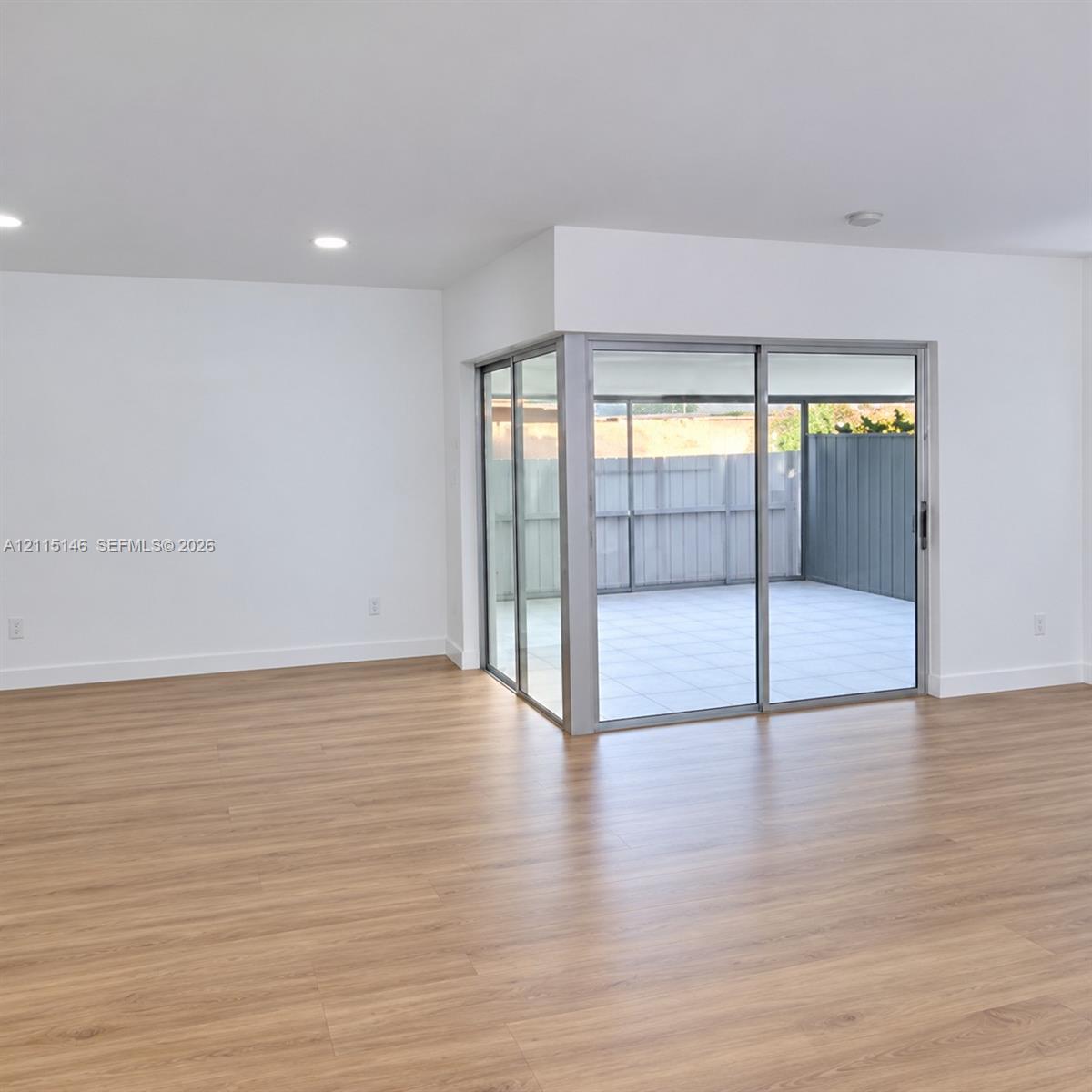 Open-concept interior featuring light wood-finish flooring, recessed lighting, and expansive corner sliding glass doors revealing a tiled patio