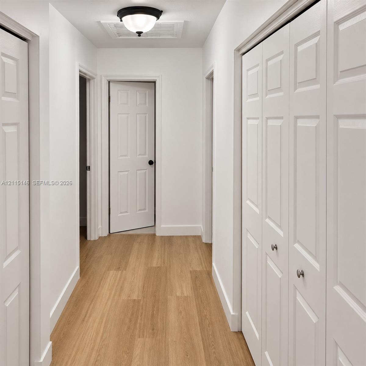 Interior hallway featuring wood-finish flooring, white six-panel doors, bi-fold closet doors with polished hardware, and a flush-mount ceiling light fixture