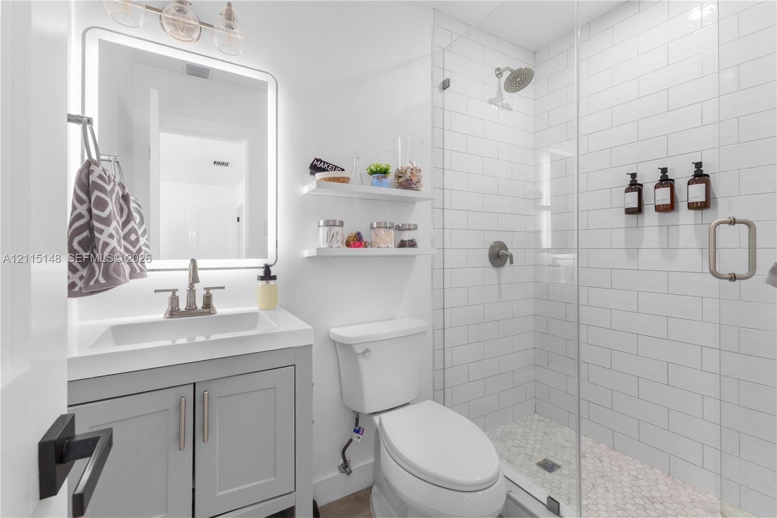 Bathroom featuring a gray shaker-style vanity with a rectangular sink, wall-mounted floating shelves, an illuminated vanity mirror, and a walk-in shower with white subway tile surround and pebble-style floor tiling