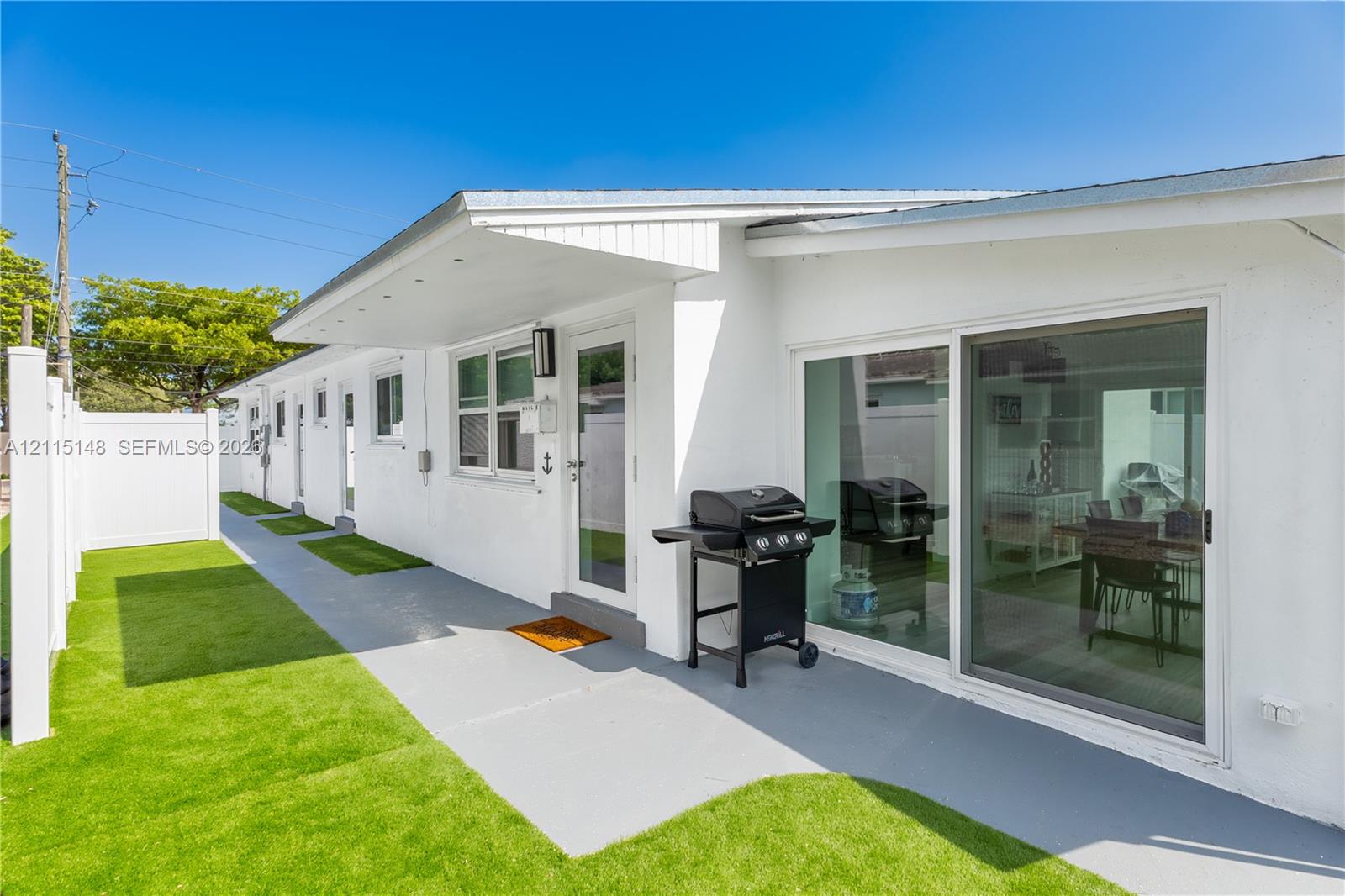 Exterior facade featuring smooth stucco siding, sliding glass doors, a covered entryway, and synthetic turf lawn bordered by a vinyl privacy fence