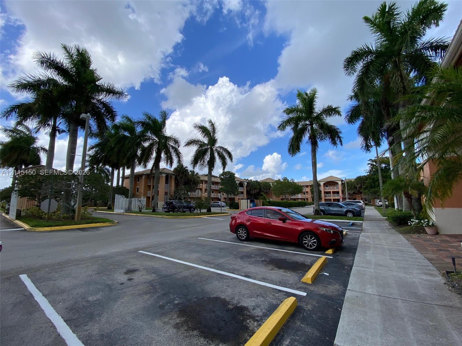 Multi-story residential complex featuring painted stucco exterior walls, paved surface parking, and mature palm tree landscaping
