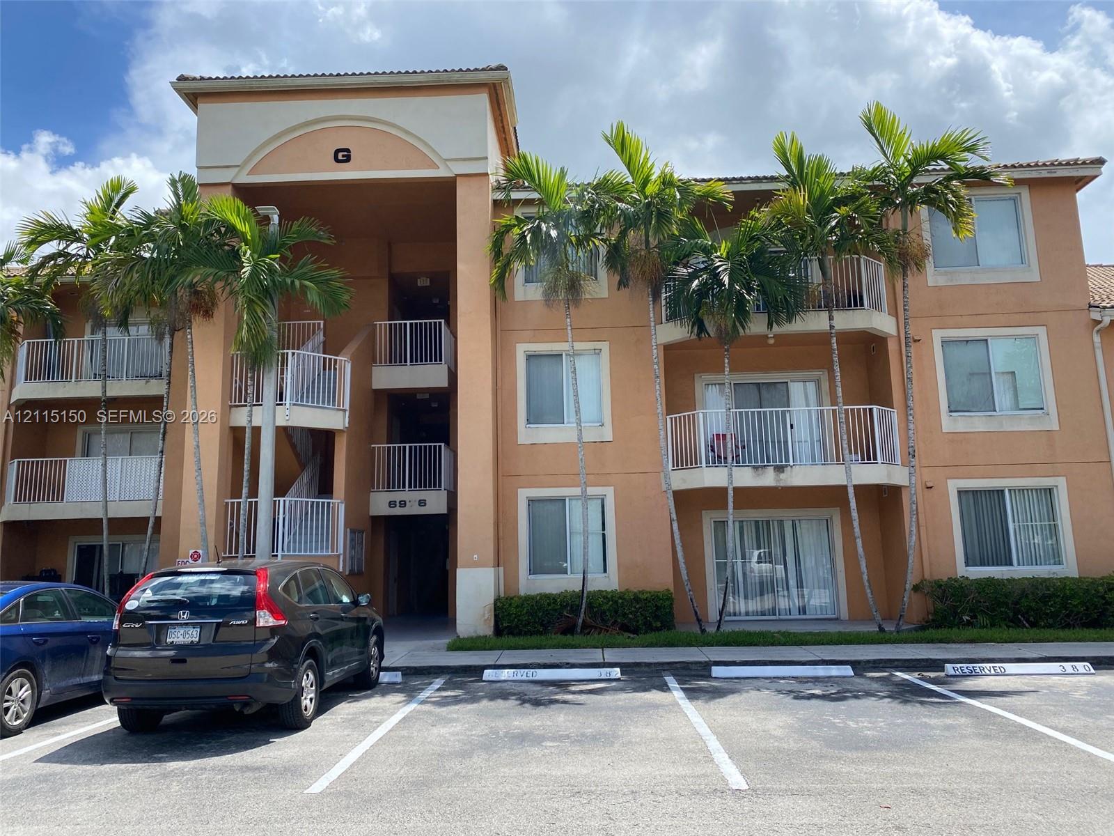 Multi-story exterior featuring a stucco facade, arched entryway, private balconies with metal railings, and mature palm trees
