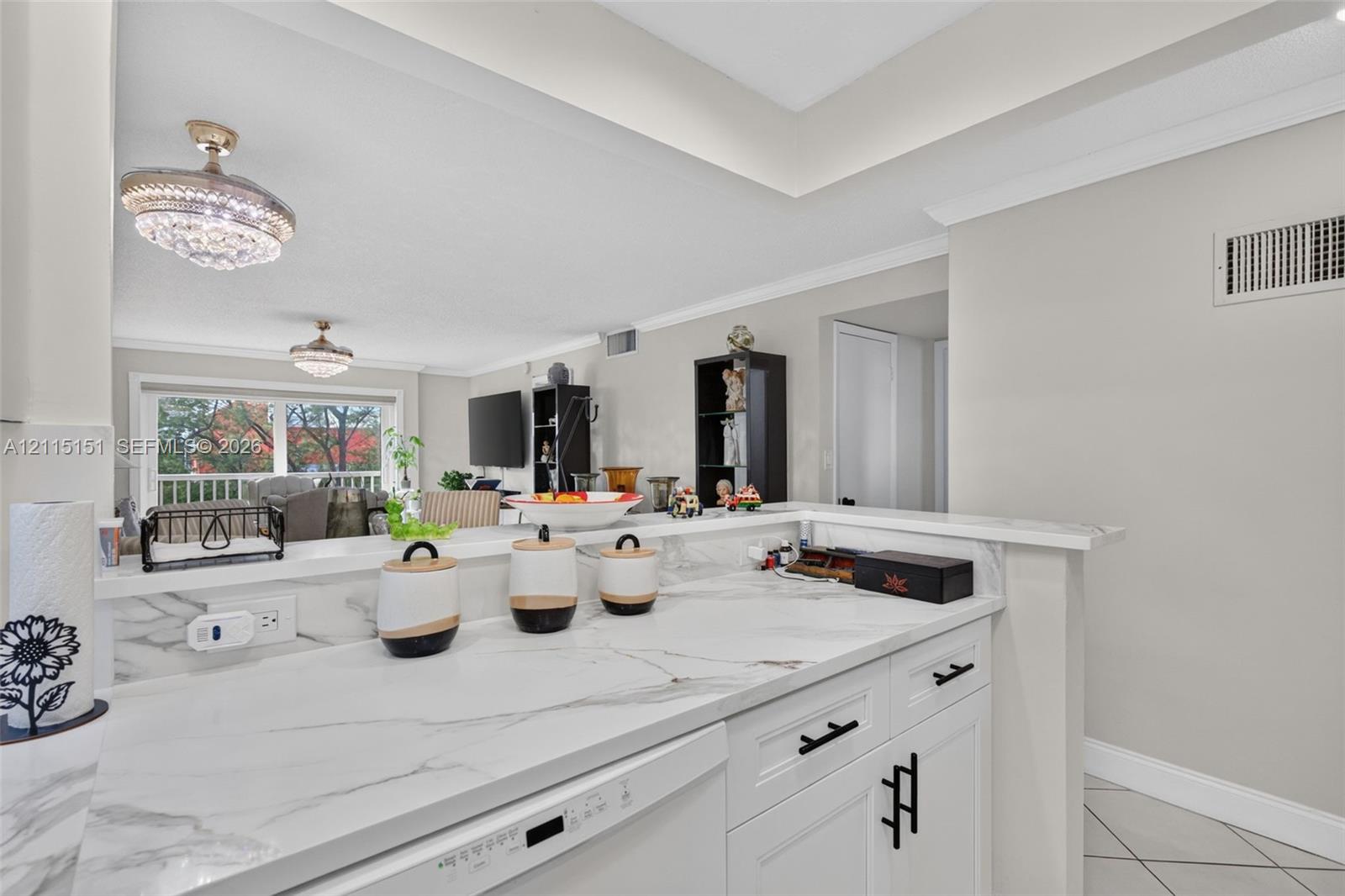 Kitchen featuring white cabinetry with matte black hardware, stone-finish countertops, ceiling crown molding, and two crystal-style pendant light fixtures Kitchen featuring white cabinetry with matte black hardware, stone-finish countertops, ceiling crown molding, and two crystal-style pendant light fixtures