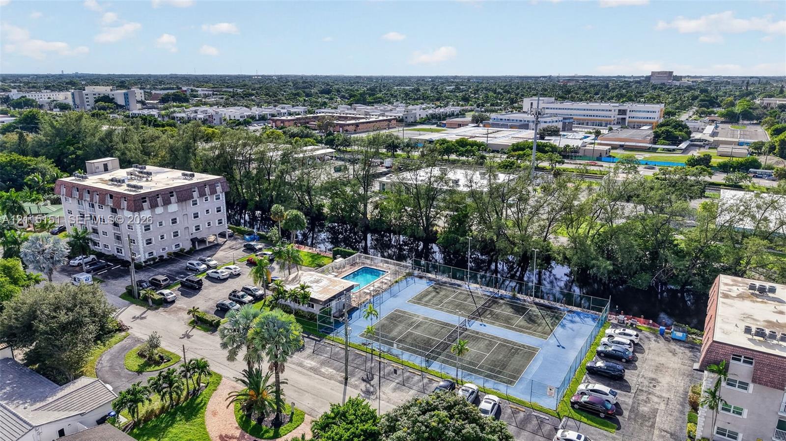 Aerial view featuring dual fenced tennis courts, an outdoor swimming pool, mature palm trees, and asphalt parking areas