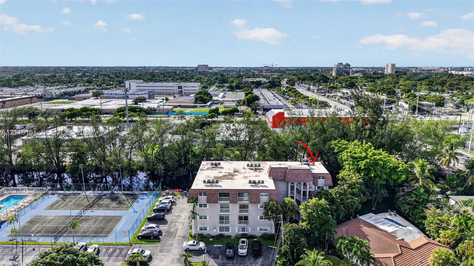 Aerial perspective of a multi-story residential building featuring a mansard roof, balconies, surface parking, and a fenced tennis court