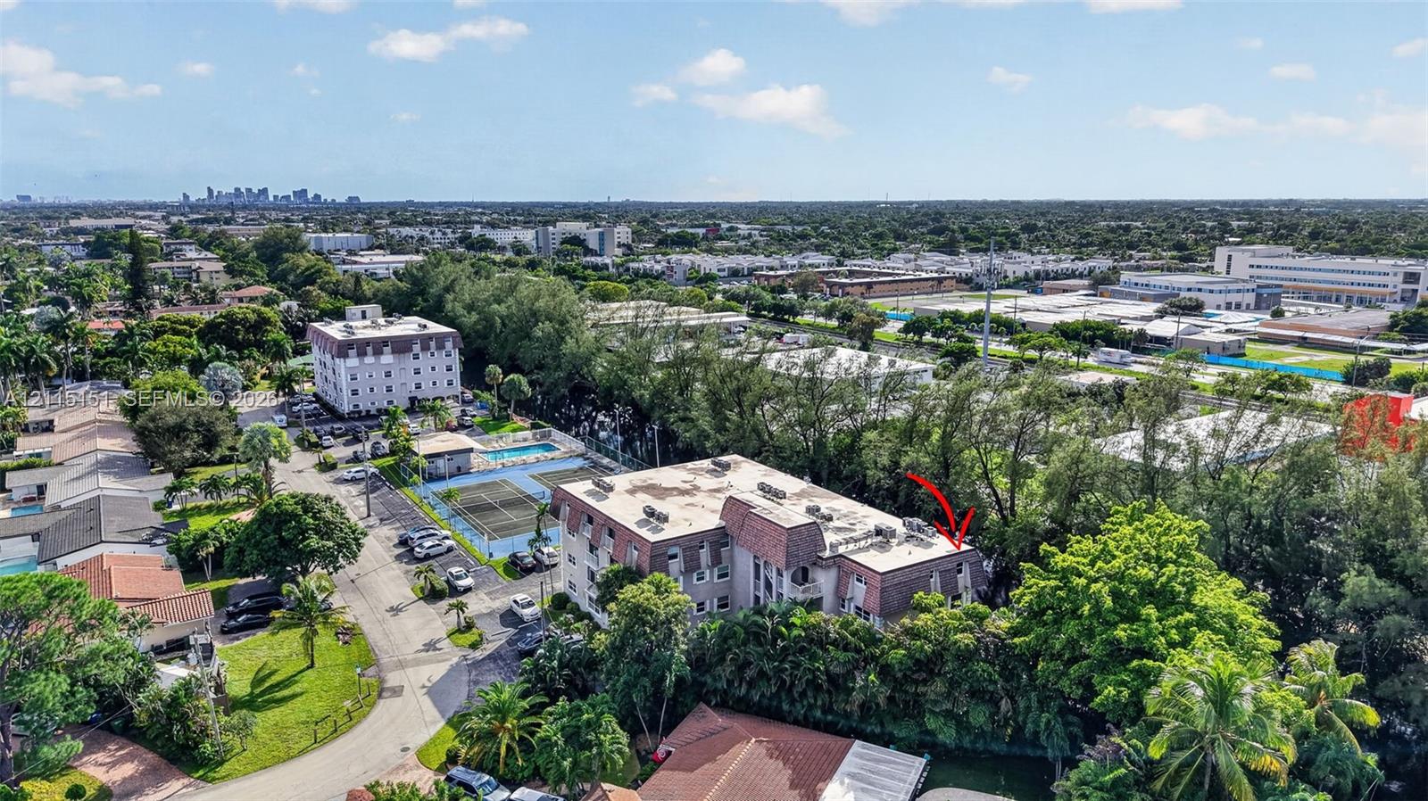 Aerial perspective of a multi-story residential building featuring a flat roofline, paved parking area, on-site tennis court, and outdoor swimming pool