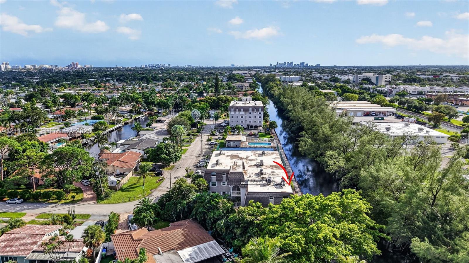 Aerial perspective of a multi-story building situated along a canal, featuring a flat roofline and dense surrounding mature landscaping