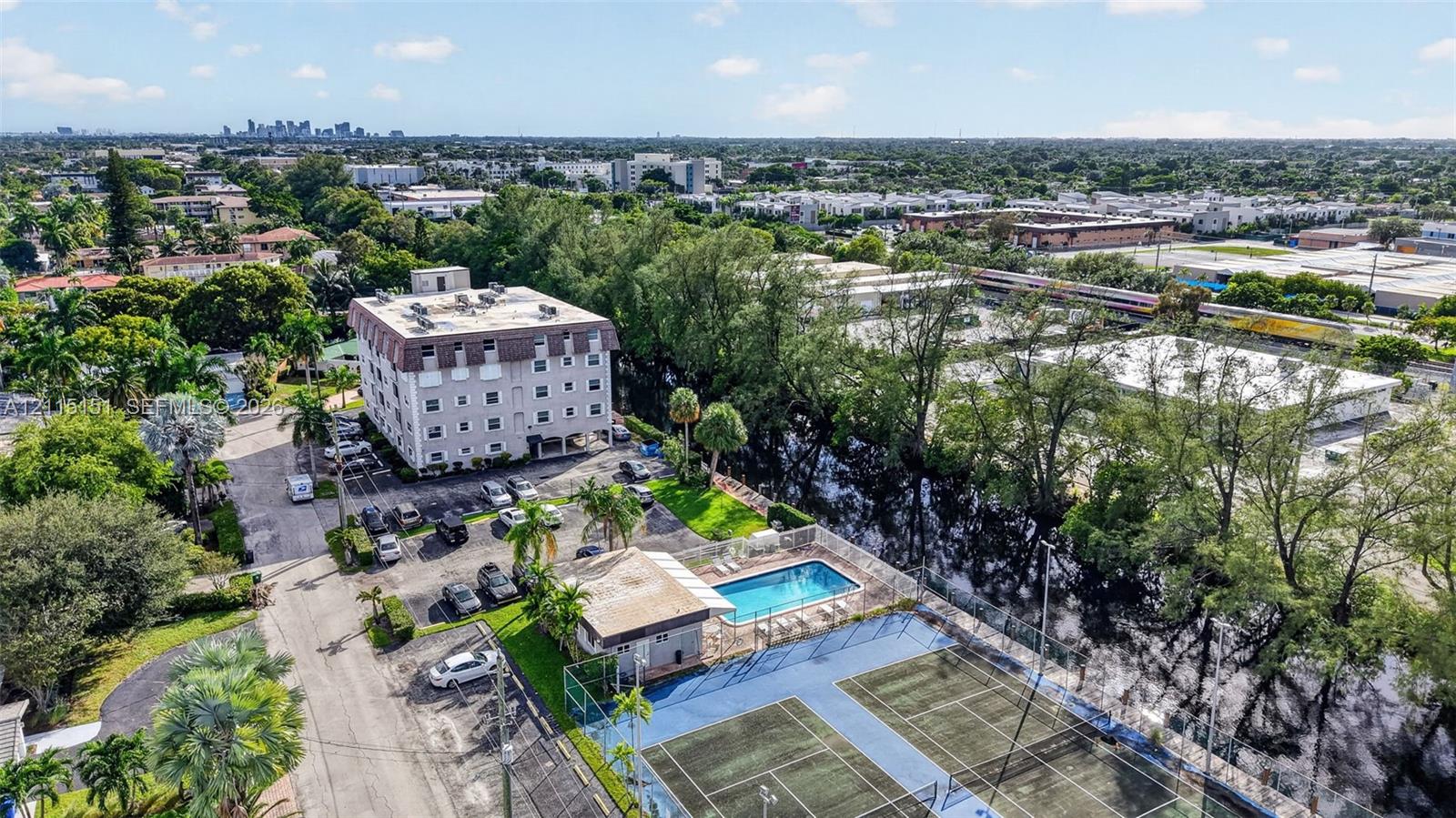 Aerial perspective of the residential complex featuring a swimming pool, two tennis courts, surface parking, and extensive mature landscaping