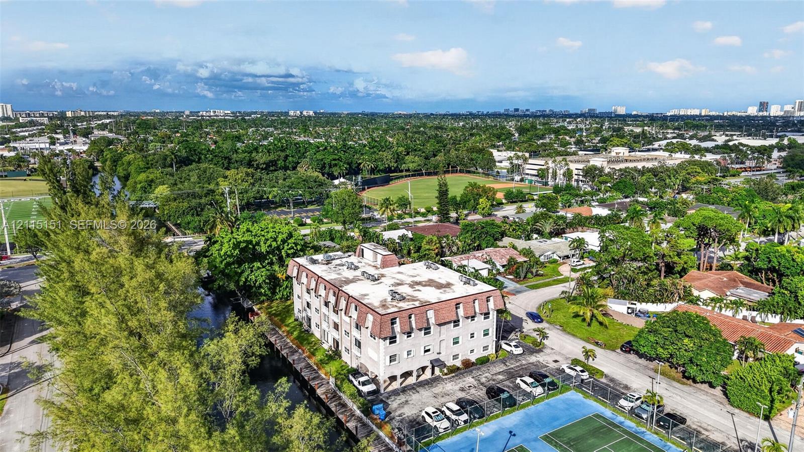 Aerial view of mid-rise multi-story building with a mansard roof, paved surface parking, and on-site tennis courts