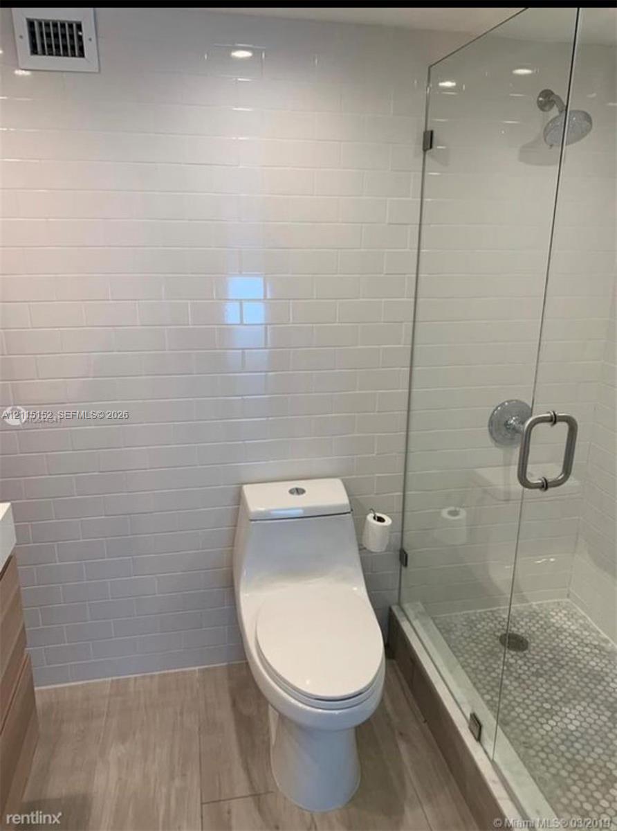 Bathroom featuring a frameless glass shower enclosure, white subway-tiled walls, a dual-flush one-piece toilet, and wood-finish flooring