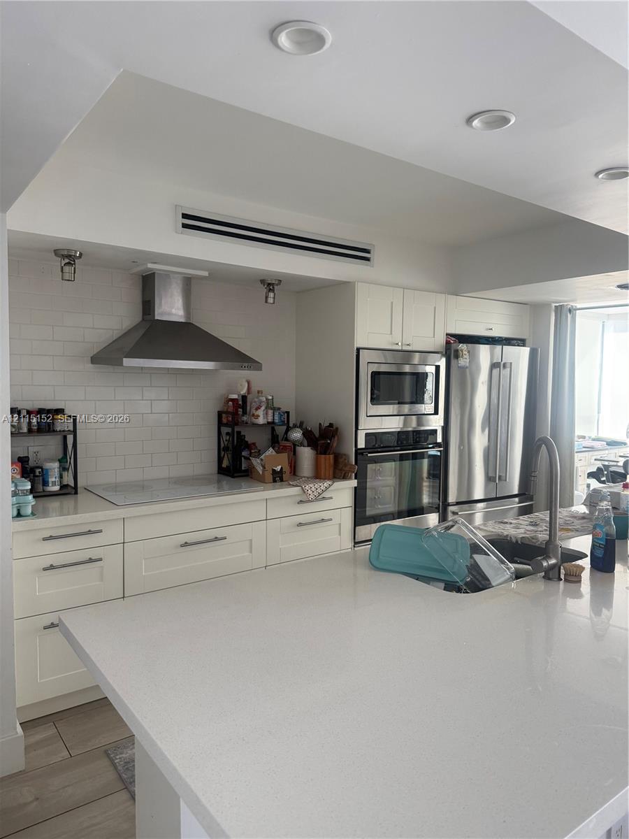 Kitchen featuring an expansive stone countertop island, custom white cabinetry, stainless steel double wall ovens, subway tile backsplash, and a stainless steel range hood