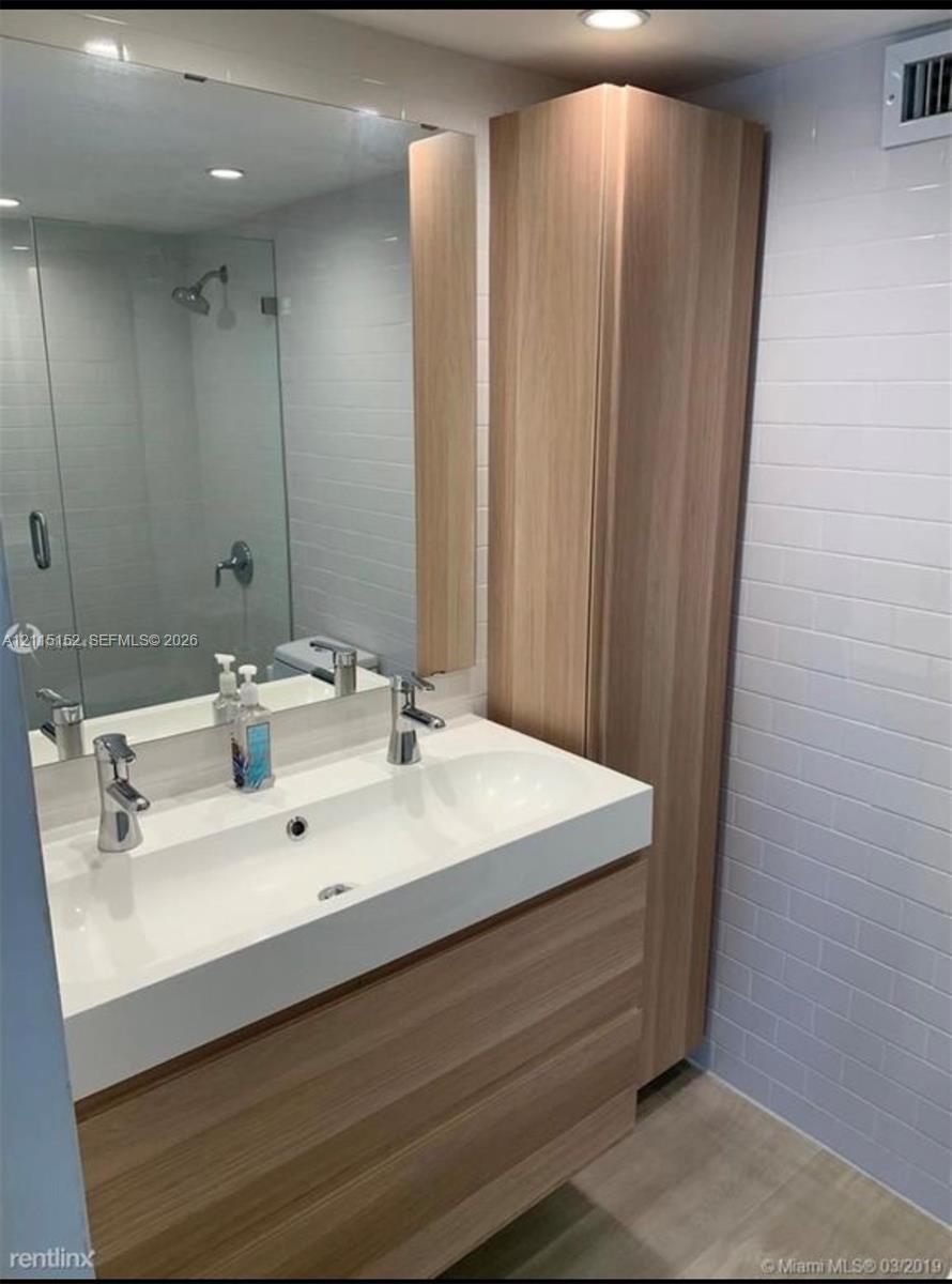 Modern bathroom featuring a wood-finish vanity with a double-sink basin, a full-height wood-finish storage cabinet, a wall-to-wall mirror, and a glass-enclosed shower with white subway tile wall cladding